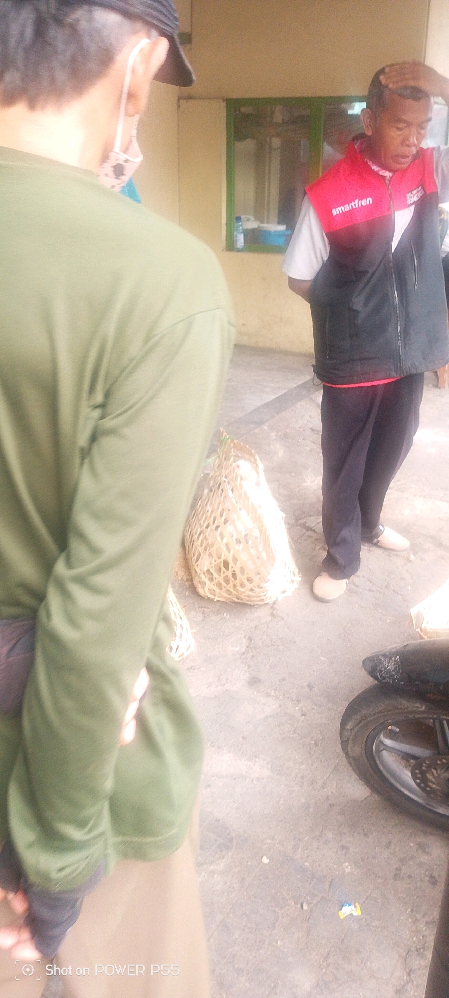 In an outdoor setting with a concrete ground, two individuals are visible. The person in the foreground, from the back, wears a green long-sleeved shirt and a black cap. They are facing away from the camera. To their right, a second individual, a man, is facing the camera. He has short hair and is wearing a red vest with "smartfren" printed on it over a white shirt, and dark pants. He is touching his head with his right hand. In front of the man, on the ground, is a woven basket containing what appears to be white poultry. To the right of the scene, the front wheel and part of a motorcycle are visible. The background consists of a building with a light-colored wall and a green-framed window. The overall impression is a casual outdoor encounter, possibly a transaction or a brief interaction.