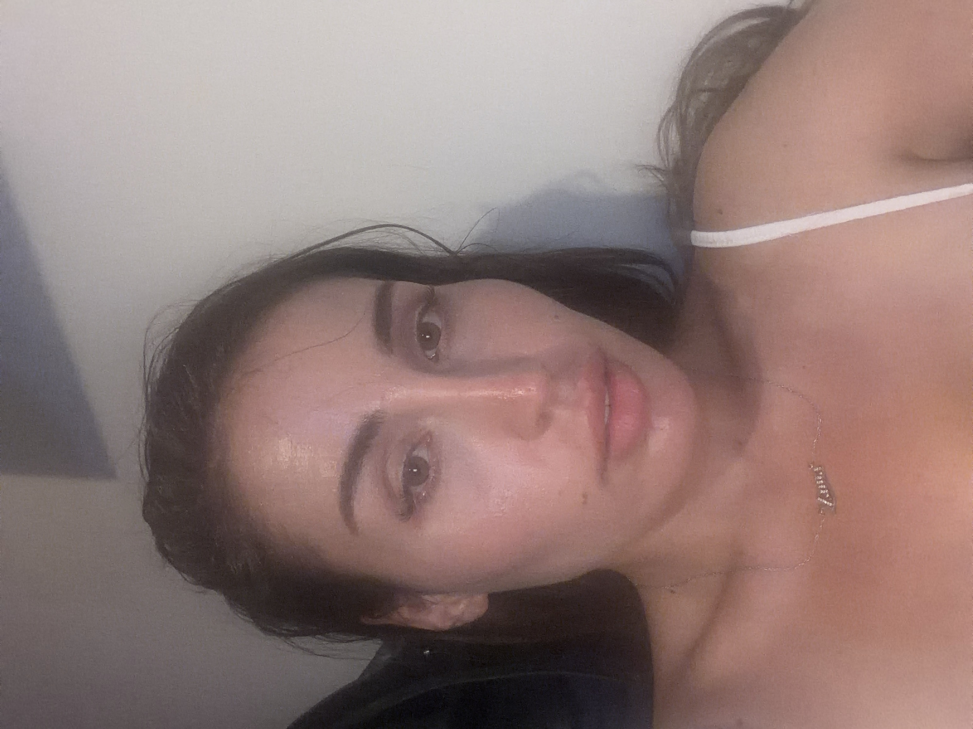 A medium shot of a woman's face is captured from a slightly low angle, with the camera focused on her upper body and face. The woman has long, dark hair that is slightly damp and appears to be pulled back loosely. She is wearing a white spaghetti strap top and a delicate silver necklace with a pendant that spells out "Zana" in cursive. Her skin is luminous and appears to have a slight sheen, possibly due to sweat or a dewy makeup finish. Her eyes are open and looking towards the camera, with visible eyelashes. Her lips are slightly parted, revealing a hint of her teeth. The background is a plain, off-white wall, with a dark shadow on the left side of the image, suggesting the presence of furniture or an object out of frame. The lighting is soft and diffuse, creating a gentle glow on her face. This image appears to be a personal photograph, likely taken indoors in a residential setting. The location is identified as Tersefanou, Cyprus.