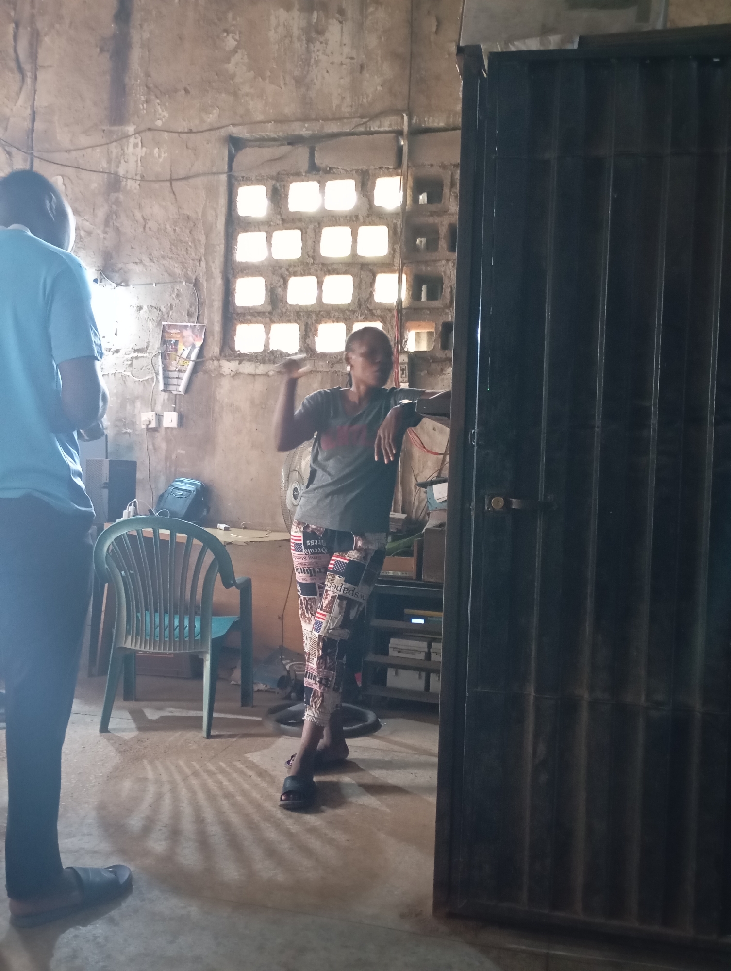 A medium shot captures two individuals in what appears to be a room in Shendam, Nigeria. On the left, a person wearing a light blue t-shirt and dark pants stands with their back partially to the camera, facing away from a tall, dark metal door that dominates the right side of the frame. The person on the left is looking towards the right. To the right of this individual and slightly further into the scene, another person stands wearing a gray t-shirt and patterned pants with red, white, and blue designs. This individual is leaning against a shelf unit and is facing towards the left, gesturing with their right arm. The environment is dimly lit with light filtering through a series of square openings in the wall behind the individuals. A green plastic chair is positioned in the foreground between the two people, and a fan is visible behind the person on the right. The walls are unpainted and appear to be made of concrete blocks.