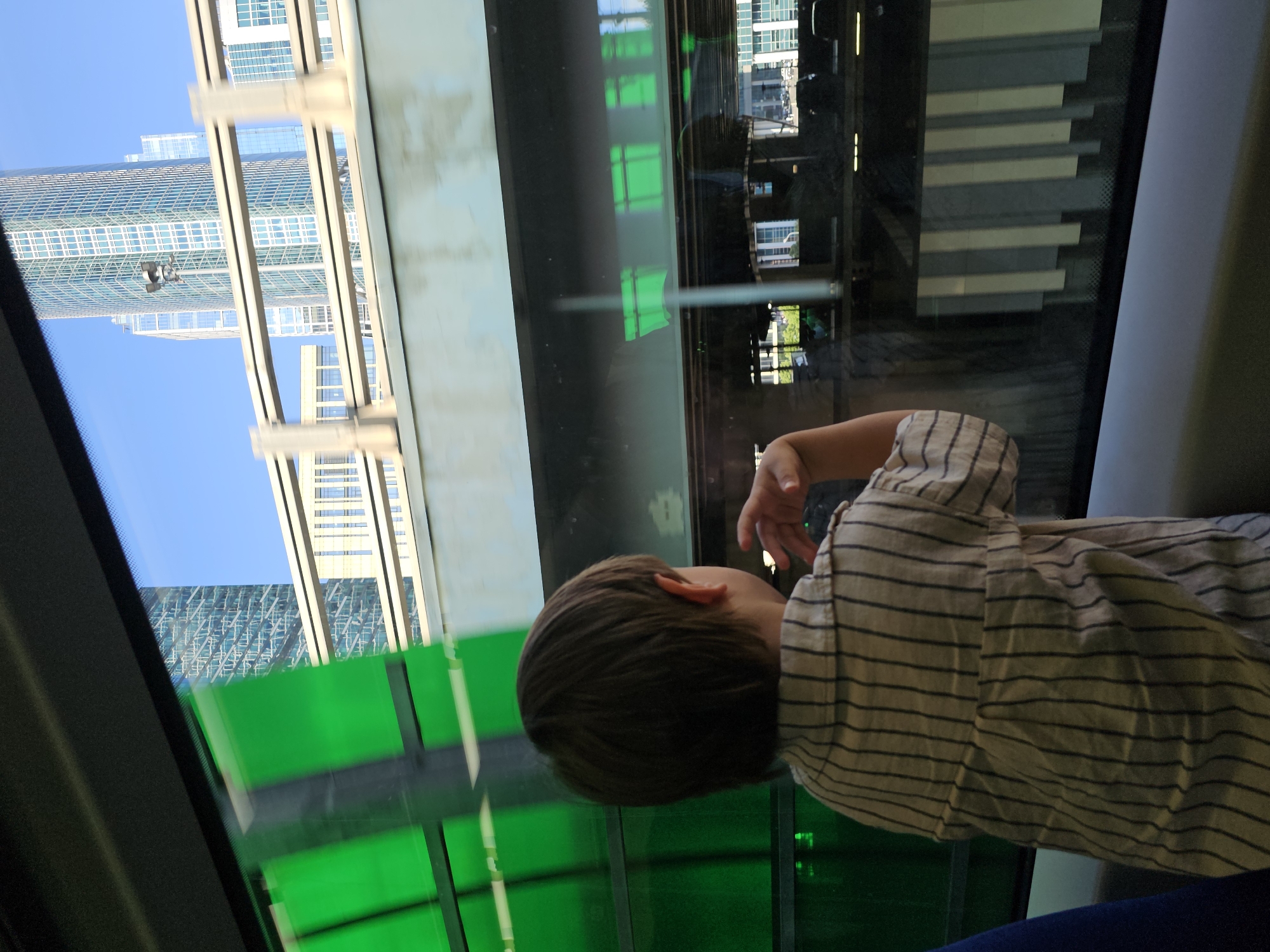 This media file depicts a young child, viewed from behind and from the side, wearing a striped shirt. The child is seated, looking out of a window. The setting appears to be a mode of transportation, possibly a train or subway, given the view through the window and the reflective surfaces.
Through the window, the child is looking at a cityscape with several modern skyscrapers. The buildings are tall and have glass facades, reflecting the clear blue sky. Some of the buildings have a geometric pattern of windows. In the foreground of the window, there are some green structures, possibly part of the transportation infrastructure or sound barriers. Reflections of lights and interior elements of the vehicle are visible on the glass.
The overall impression is of a daytime scene, likely late morning or early afternoon, given the brightness of the sky and the lighting on the buildings. There are no clear indications of weather, other than the clear sky. The child's posture and focus on the outside suggest curiosity or observation. The scene evokes a sense of travel through a contemporary urban environment.
There is no visible text in the image. The crops provide closer views of the buildings, the child's clothing, and the reflections in the window, confirming the urban setting and the details of the child's attire and the surrounding environment. The Moscow, Russia location context suggests this is likely a view of the Moscow skyline.