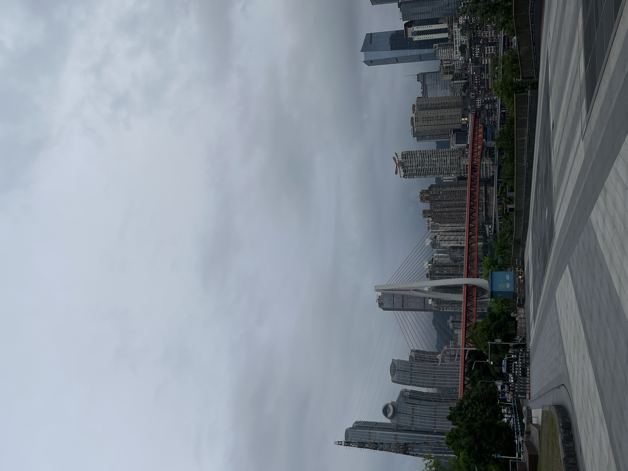 The image shows a cityscape of Jiangbeicheng Subdistrict, China, taken from a high vantage point. A red and white cable-stayed bridge spans a river, surrounded by densely packed high-rise buildings. The sky is overcast, with grey clouds dominating the scene. A paved walkway with a striped pattern is visible in the foreground. Trees and other greenery are interspersed among the buildings.