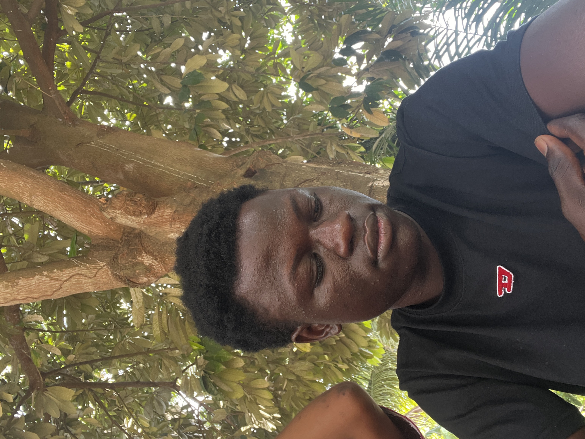 The image shows a person with dark skin, wearing a black t-shirt with a red "R" emblem, lying under a tree. The person's eyes are closed. The background shows branches and leaves of the tree, with sunlight filtering through. Based on the prompt, the location is Badagry, Nigeria.