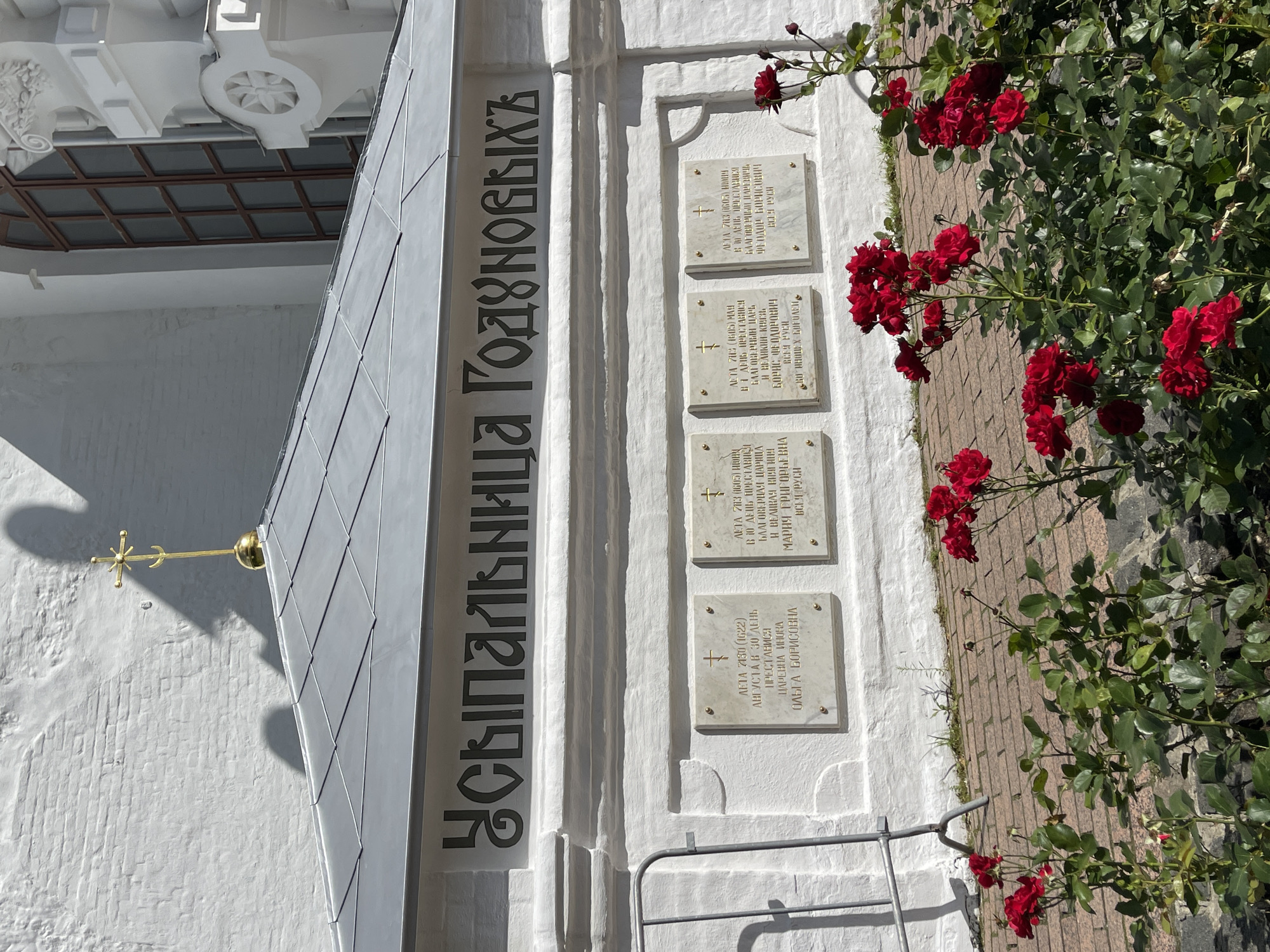 The image depicts a section of the Trinity Lavra of St. Sergius in Sergiev Posad, Russia. The focal point is a white brick wall with a grey, pitched roof extending over it. Above the wall, large, dark, stylized Cyrillic lettering reads "УСЫПАЛЬНИЦА ГОДУНОВЫХ," which translates to "Tomb of the Godunovs." Below this inscription, four white plaques with gold crosses are affixed to the wall. Each plaque contains Russian text, likely commemorating individuals buried in the tomb. Red roses are blooming in the foreground, with their bushes and leaves obscuring the lower portion of the wall and a brick pathway. A metal fence is visible on the left side of the image. The architecture suggests a historical religious site.