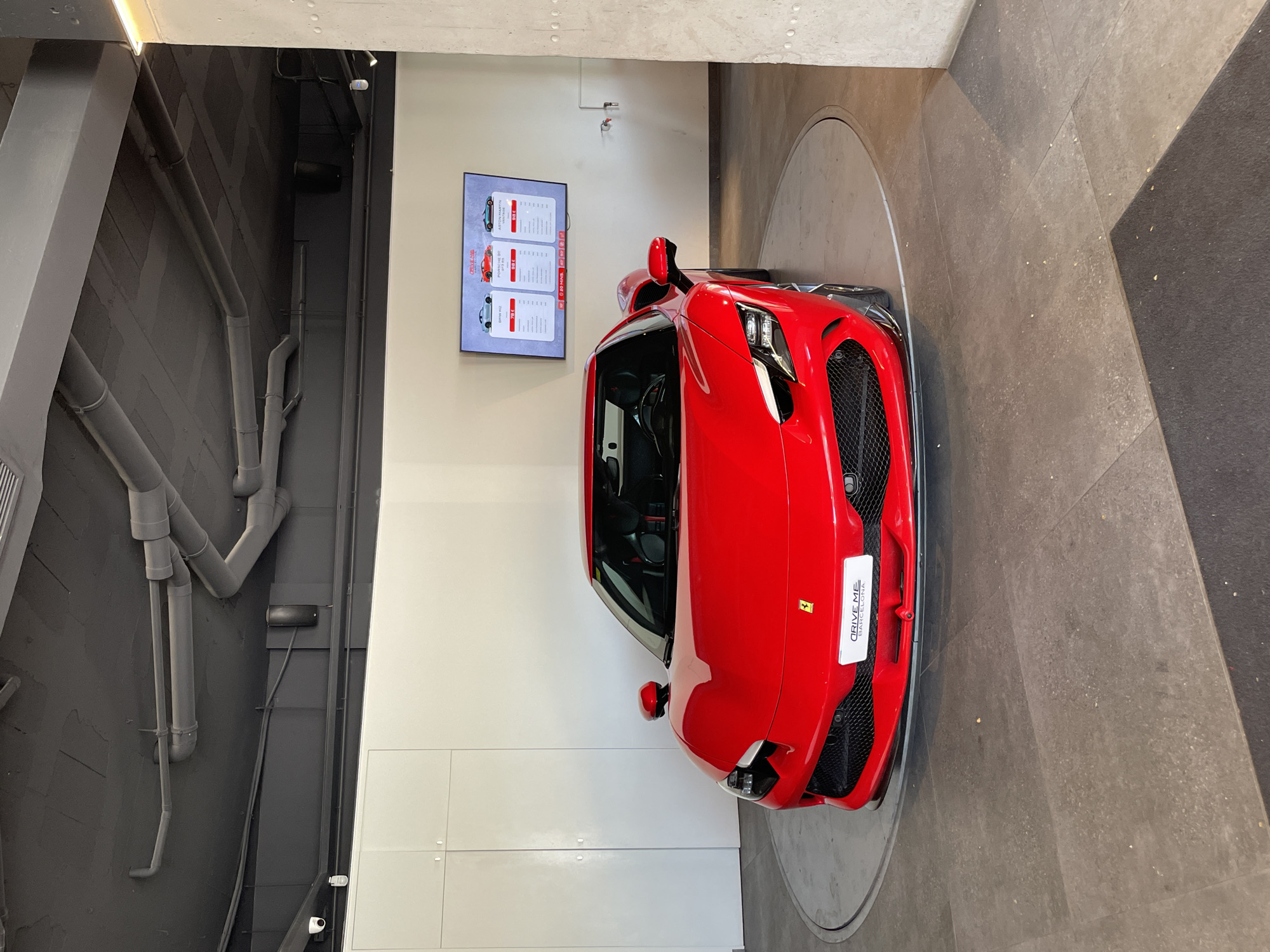 A vibrant red Ferrari F8 Tributo is the central subject of this high-angle indoor shot, displayed on a circular rotating platform. The car's sleek front end, featuring sharp headlights, a prominent black grille, and air intakes, is clearly visible. A white front license plate holder on the car reads "DRIVE ME BARCELONA" next to a small Ferrari logo.
The setting is a modern showroom or car experience center, exhibiting an industrial aesthetic. The ceiling reveals exposed concrete structures, integrated lighting, and a network of gray pipes and ducts. The walls are a combination of clean white panels on the right and a textured dark gray surface with subtle patterns and additional exposed piping on the left. The floor surrounding the car is laid with large tiles in varying shades of gray and brown, forming geometric patterns.
On the white wall, a digital display screen advertises car rental services under the title "DRIVE THE CARS OF YOUR DREAMS!". The screen lists several luxury vehicles and their rental prices for specific distances: a "PORSCHE 992" for "50 KM / 112 €", the "FERRARI F8 TRIBUTO" for "50 KM / 204 €", and an "ASTON MARTIN VANTAGE" for "50 KM / 111 €". Additional options for "20 KM / 58 €" and "40 KM / 92 €" are also presented.
No individuals are present in the scene. The overall impression conveyed is that of an exclusive car rental or driving experience facility, confirmed by the "DRIVE ME BARCELONA" text, aligning with the provided location context of Barcelona, Spain. The environment is well-lit, typical of an indoor commercial space.