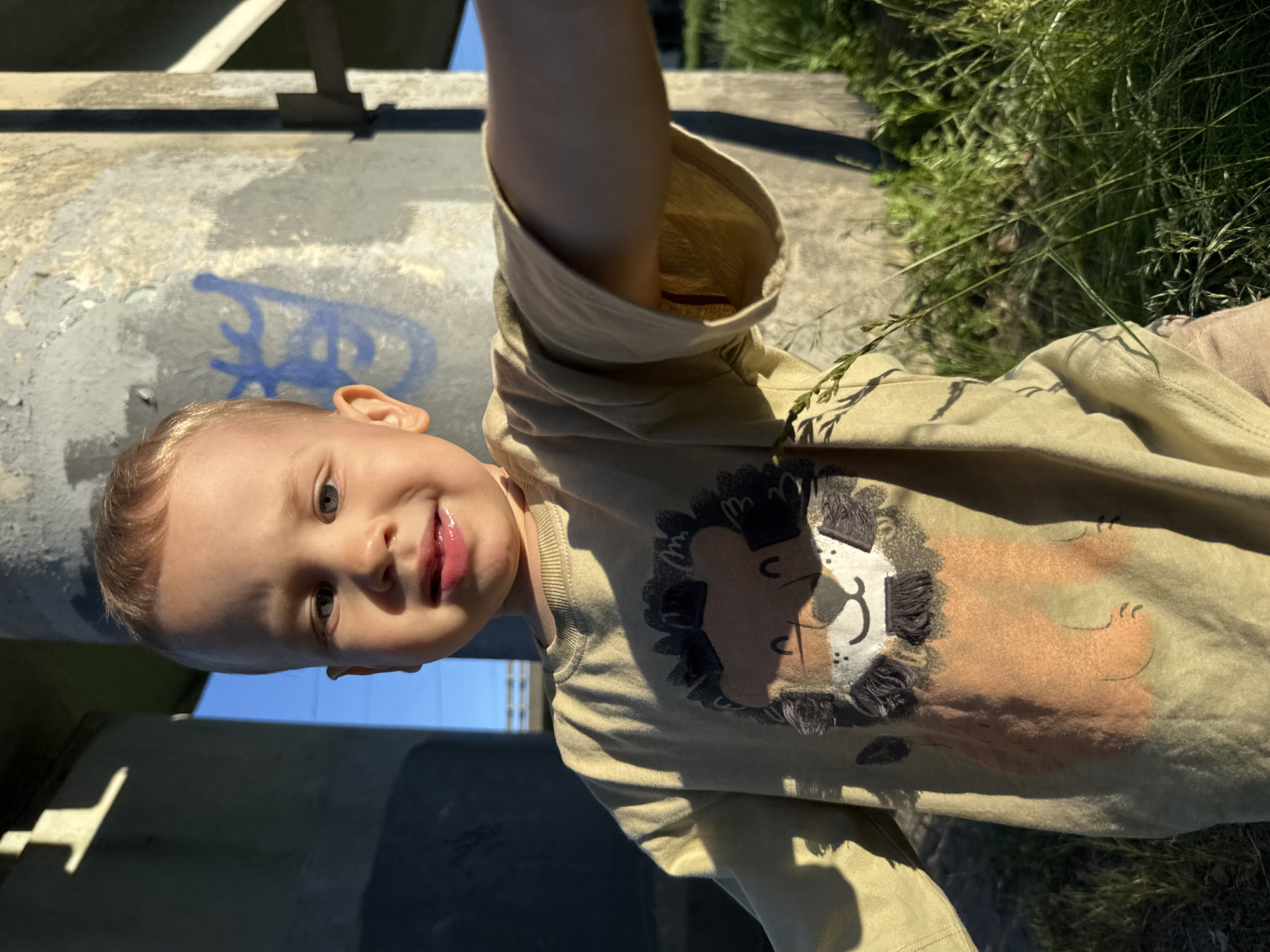 A young, fair-skinned child with blonde hair and blue eyes is depicted in a front-facing, close-up photograph. The child looks directly at the camera with a slight smile, and their lips appear moist. They are wearing a light olive-green long-sleeved shirt featuring a large, stylized cartoon lion graphic on the chest. The child's right arm is extended forward, suggesting a selfie or close-held camera perspective. Light-colored pants are partially visible in the lower right. The scene is an outdoor location in Rusanovo, Russia, set beneath a concrete overpass or bridge structure. Two weathered concrete support pillars are visible in the background, with faded blue graffiti present on the right-hand pillar. Strong sunlight illuminates the child and immediate environment, casting distinct shadows. Green grass and natural foliage are visible in the foreground and bottom right, indicating an overgrown area beneath the structure. Segments of blue sky are visible through the structure's openings.