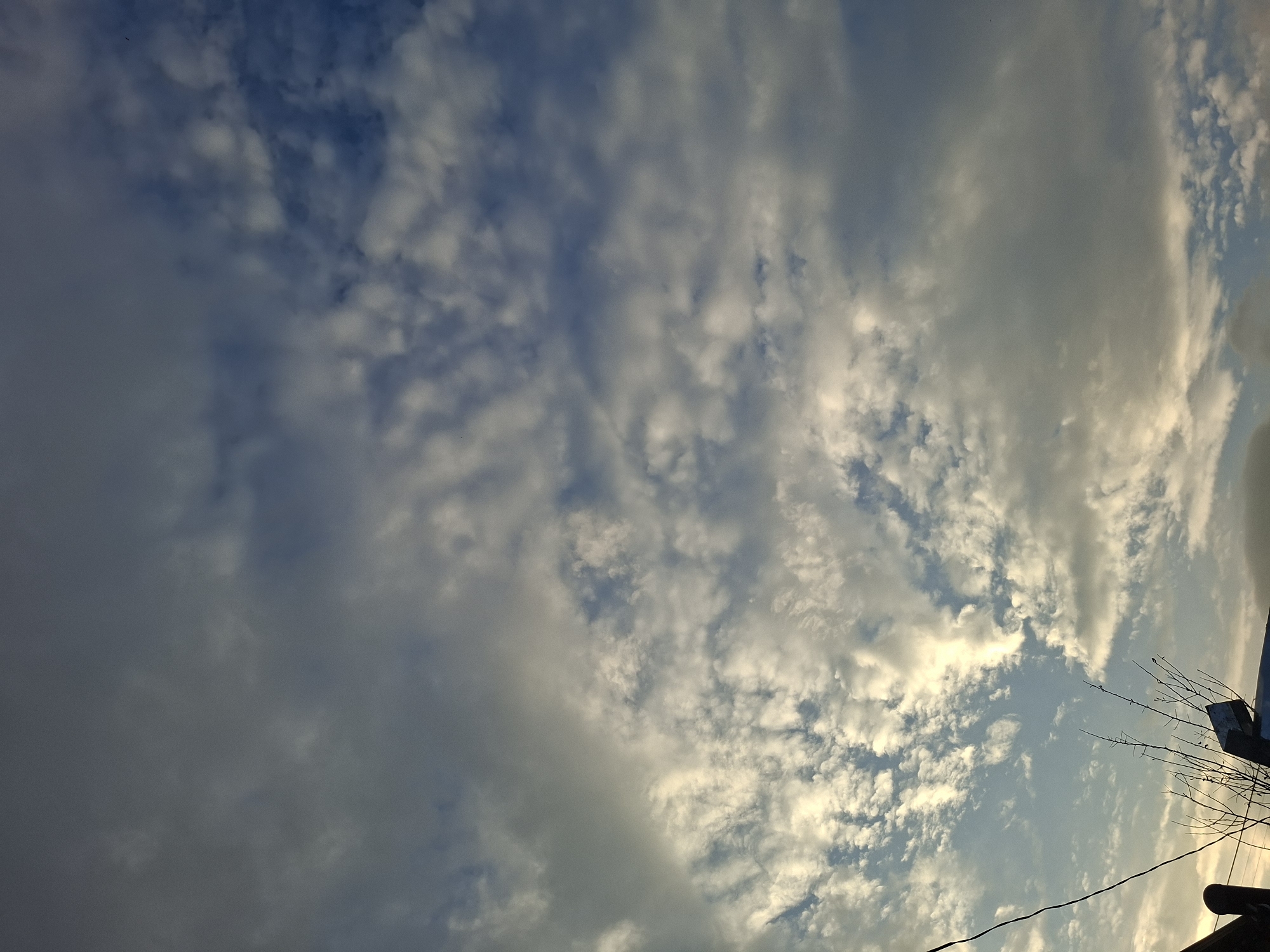 The image shows a sky with scattered clouds of varying thickness and texture, illuminated by sunlight. The clouds range from wispy and translucent to dense and opaque, with patches of blue sky visible between them. The lighting suggests it is either early morning or late afternoon. In the lower left corner of the frame, a portion of a dark pole or fence is visible, along with the silhouette of a few dry branches. The image is a natural scene, likely taken outdoors. No specific location in Tbilisi, Georgia is identifiable from the visual information presented.