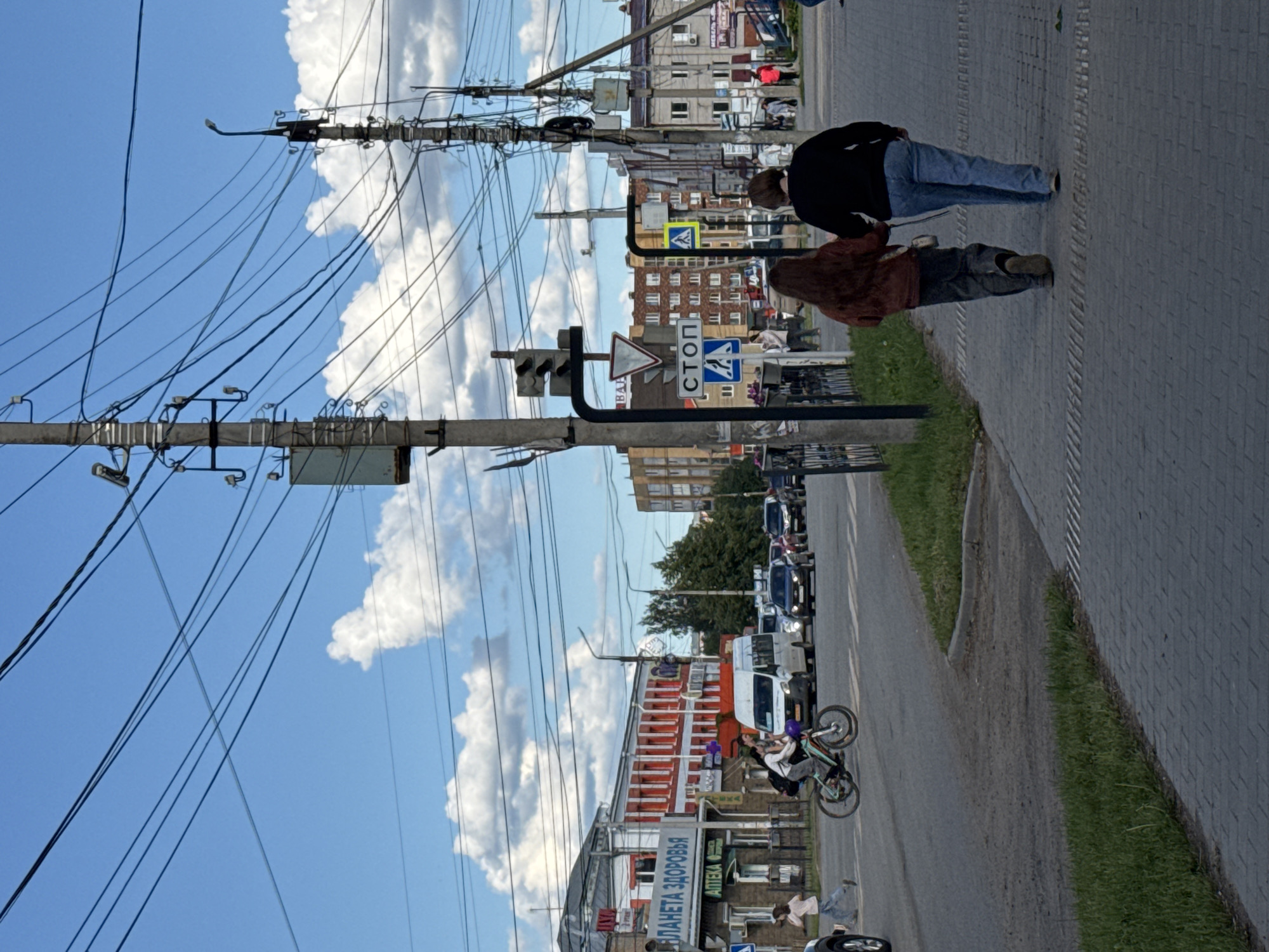 The image shows a street scene in Mozhga, Russia. Power lines dominate the upper left part of the frame against a backdrop of blue sky with cumulus clouds. A stop sign is visible. Two people stand on the sidewalk on the right. Further down the street, a bicyclist performs a wheelie. Cars are parked along the roadside. Buildings line the street. A sign with Russian text is visible on one building, bearing the words "ПЛАНЕТА ЗДОРОВЬЯ" (PLANET OF HEALTH).