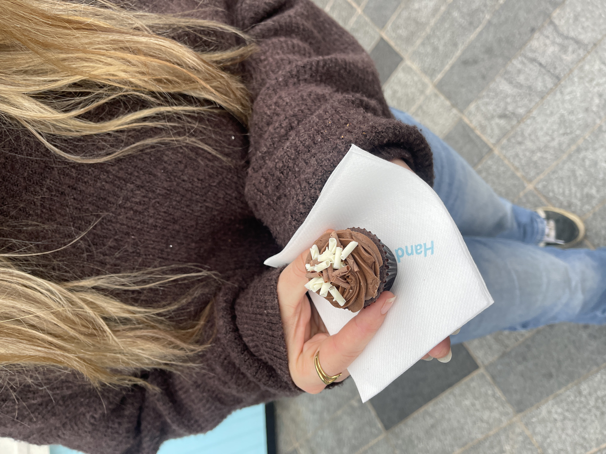 The image, taken from a top-down perspective, captures a person holding a chocolate cupcake outdoors in what appears to be London, United Kingdom. The subject's upper body, right arm, and legs are visible. They have long, light blonde hair cascading over a textured, dark brown knitted sweater. A gold-colored ring with a double band design is worn on their right hand's ring finger.
The person is holding a small, dark chocolate cupcake with a generous swirl of chocolate frosting, topped with white chocolate shavings. The cupcake rests on a white paper napkin, which is held between their thumb and index finger. The word "Hand" is visible in light blue text on the napkin, oriented upside down from the viewer's perspective.
Below the sweater, light blue jeans are visible, suggesting a casual attire. The background consists of a grey tiled pavement with distinct grout lines, typical of urban outdoor spaces. Part of a dark shoe or trainer is also visible in the bottom right corner, suggesting the person is standing or walking. The lighting appears natural and bright, indicating it is daytime.