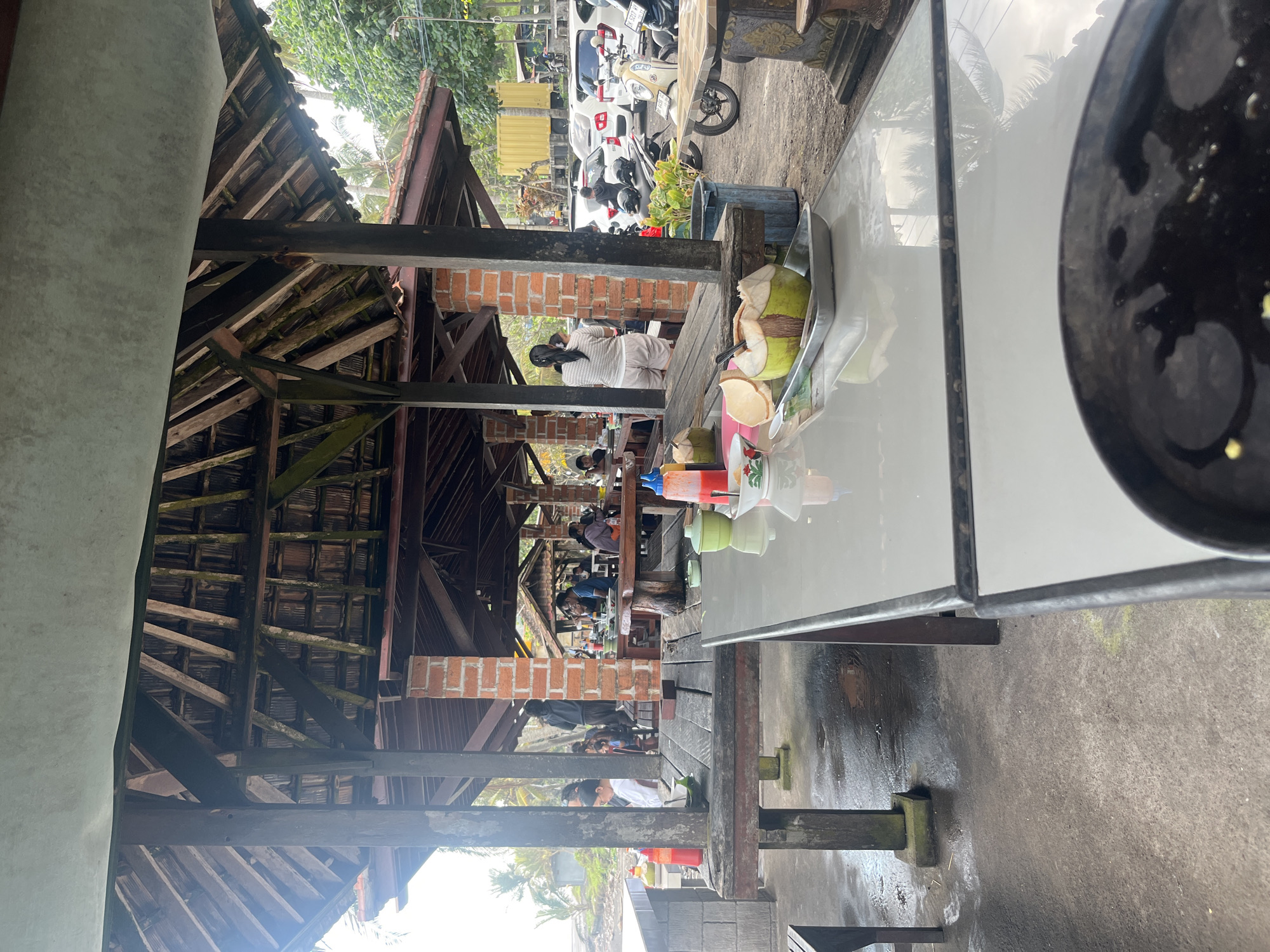 This image shows an outdoor seating area at what appears to be a casual eatery or restaurant, possibly in Pekutatan, Indonesia. The setting is characterized by a rustic, open-air structure with a wooden roof and supporting beams, suggesting a tropical environment. Several people are seated at tables, engaged in conversation or eating.
In the foreground, a glass-topped table is visible, with a tray holding a young coconut and some cut coconut pieces. The table surface reflects the sky and surrounding greenery, hinting at daylight hours. Beyond the table, the covered seating area stretches back, with more tables and patrons. The architecture features exposed wooden beams and terracotta brickwork.
In the background, several motorcycles and a white car are parked, indicating this might be a place accessible by road. Lush green foliage can be seen, further suggesting a warm, possibly humid climate. The overall atmosphere appears relaxed and informal. There is no discernible text in the image.