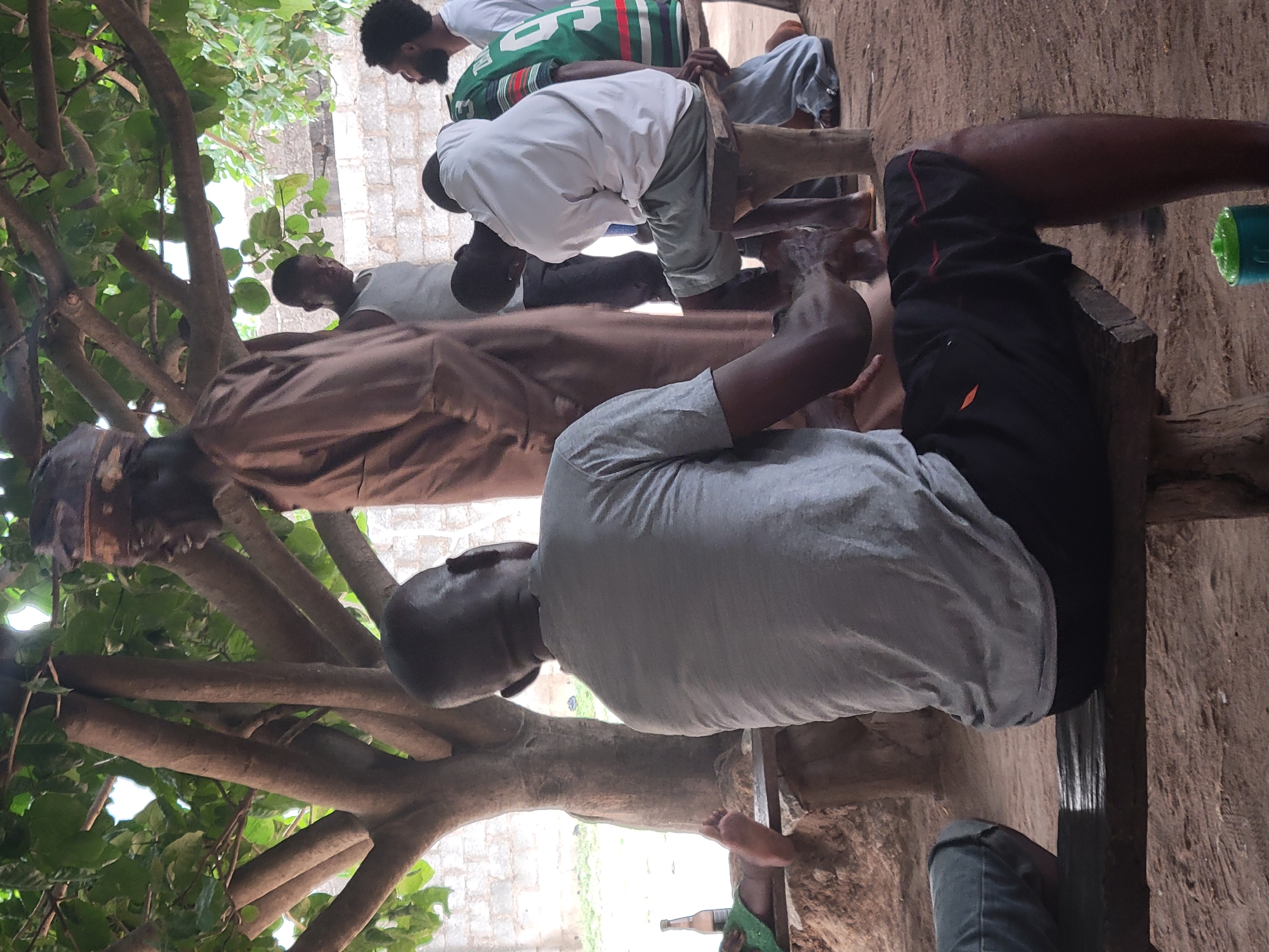 A group of men is sitting and standing outdoors, likely in Madalla, Nigeria. The scene features men of varying ages. One man wears a brown robe and a skullcap. Another man wears a gray shirt and black shorts. A man further back in the group wears a white shirt. Another man is wearing a green and white jersey. They are positioned around a wooden bench. A tree with green leaves is visible in the foreground. The ground is dirt.