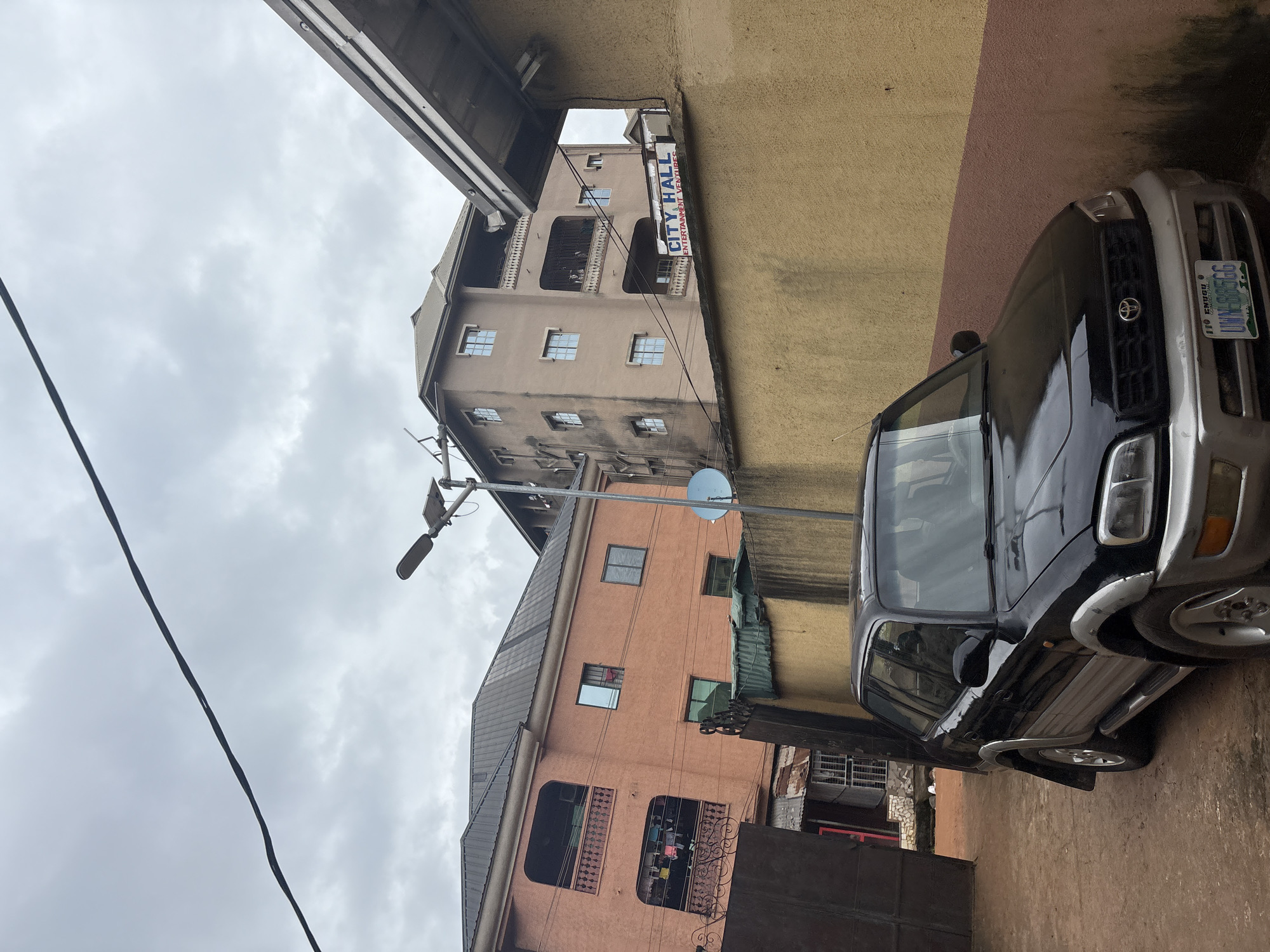 The image depicts a street scene in Onitsha, Nigeria, under an overcast sky. In the foreground, a black Toyota RAV4 is parked, facing left. The vehicle has a two-tone paint scheme, with a silver lower section and black upper section. Its license plate is visible and reads "110 ENUGU UWW 885GG".
Behind the car, a beige wall extends across the frame. To the left of the wall, a tall, orange building with balconies and laundry hanging from them is visible. To the right of the orange building, a taller, light brown building with multiple windows and balconies stands. A sign on this building reads "CITY HALL ENTERTAINMENT VENTURES".
Further to the left, a dark metal gate or fence partially obscures a view of another building with corrugated metal roofing. A street light with a solar panel is positioned between the orange and light brown buildings, and a satellite dish is mounted on the light brown building. The ground appears to be dirt or gravel. The overall impression is of a dense urban environment.