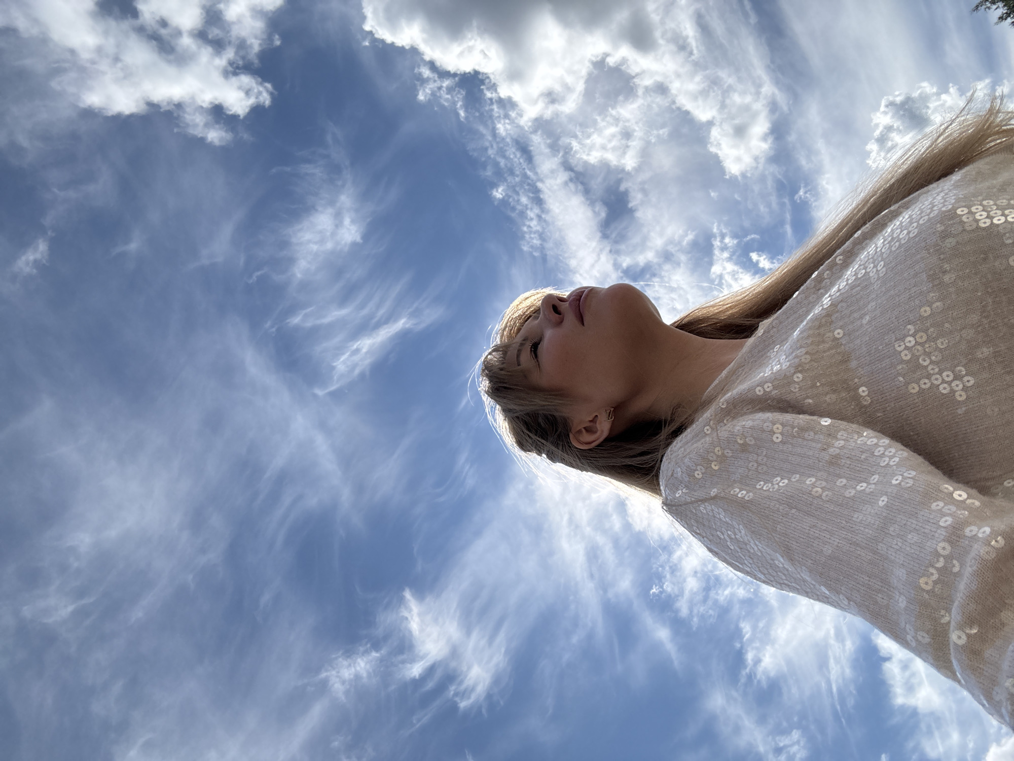 This is a low-angle, portrait-oriented, eye-level media file taken outdoors, likely in Yekaterinburg, Russia. The primary subject is a young woman, her head and upper torso visible, looking upwards. She has long, blonde hair and is wearing a cream-colored, sequined sweater. An earring with a small, gold 'V' or 'Z' shape is visible on her right ear. Her expression appears serene or contemplative as she gazes towards the sky.
The setting is a bright, clear blue sky filled with wispy, cirrus clouds that stretch across the frame. Some larger, puffier clouds are also present, particularly in the upper right quadrant. The lighting suggests it is daytime, possibly late morning or early afternoon, given the brightness and the way the light illuminates the clouds. The overall mood is peaceful and expansive, with the vastness of the sky dominating the scene. There are no visible text elements in the media file.