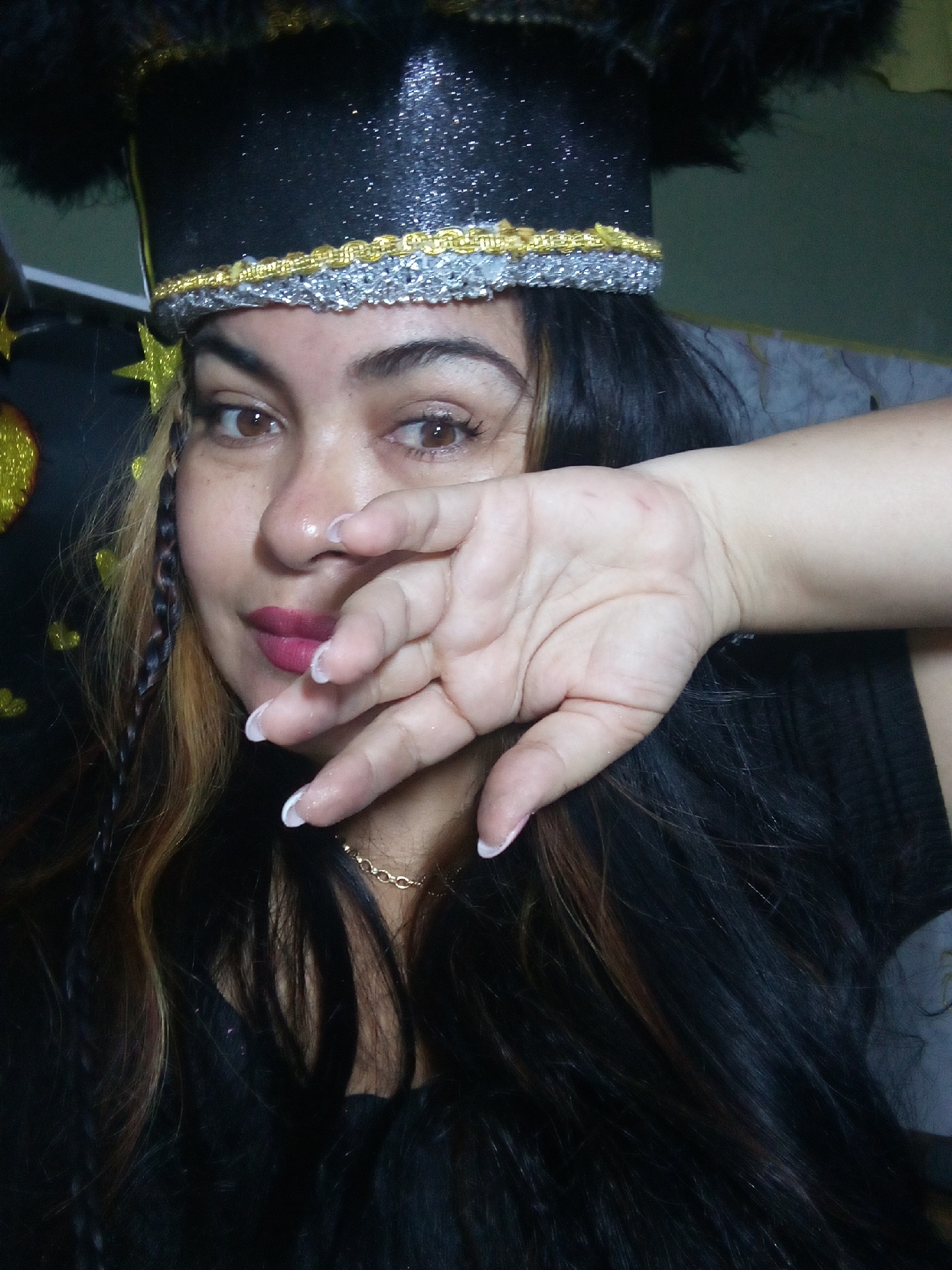 A woman is seen from the chest up. She has dark hair with blonde streaks and a braid on the left side of her face. She has light brown eyes, and her lips are painted a dark pink color. Her nails are light pink with white tips. She is wearing a black hat with gold and silver trim and black feather-like adornments on top. She is covering half of her face with her right hand. A gold chain is visible around her neck. The background includes a grey and black patterned surface and items with gold star shapes. It is not possible to determine the location as Juan Griego, Venezuela from the image.