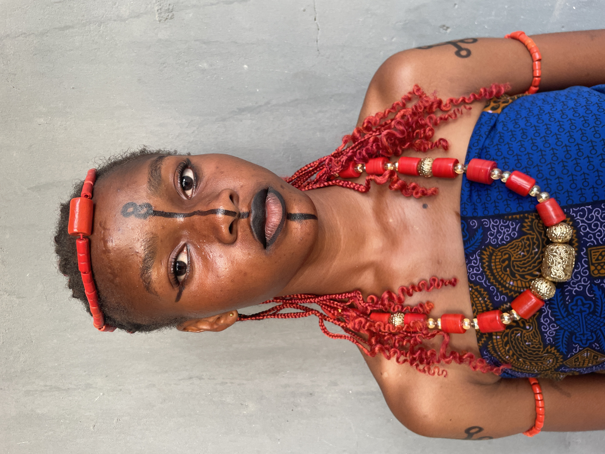 A close-up, vertically oriented portrait features a person with dark skin, adorned in what appears to be traditional or culturally inspired attire and elaborate facial markings, against a plain, light grey, textured background. The individual has short, dark, curly hair, and a red beaded headband is worn across the forehead.
The face is dramatically painted with black lines: a thick line across the forehead, another line extending from the outer corner of the left eye down the cheek, and a vertical line bisecting the dark-painted lips. The eyes are wide open and gaze slightly upwards, conveying a direct and composed expression.
Elaborate red beadwork is a prominent feature: a substantial necklace with large, cylindrical red beads interspersed with ornate gold-colored spherical and cylindrical beads. Multiple strands of curly red material, possibly hair extensions or decorative elements, are adorned with small red and gold beads, emanating from behind the ears and extending down the shoulders. A simple red beaded bracelet is on the right arm, and a dark, stylized tattoo is visible on the left upper arm. The person wears a vibrant blue fabric garment featuring intricate yellow and black patterns, suggesting traditional Nigerian textile.
The setting is understated, likely an indoor or sheltered outdoor environment in Badagry, Nigeria, with even lighting that highlights the subject's features and adornments. No specific activity beyond posing for the portrait is depicted. There is no visible text in the image.