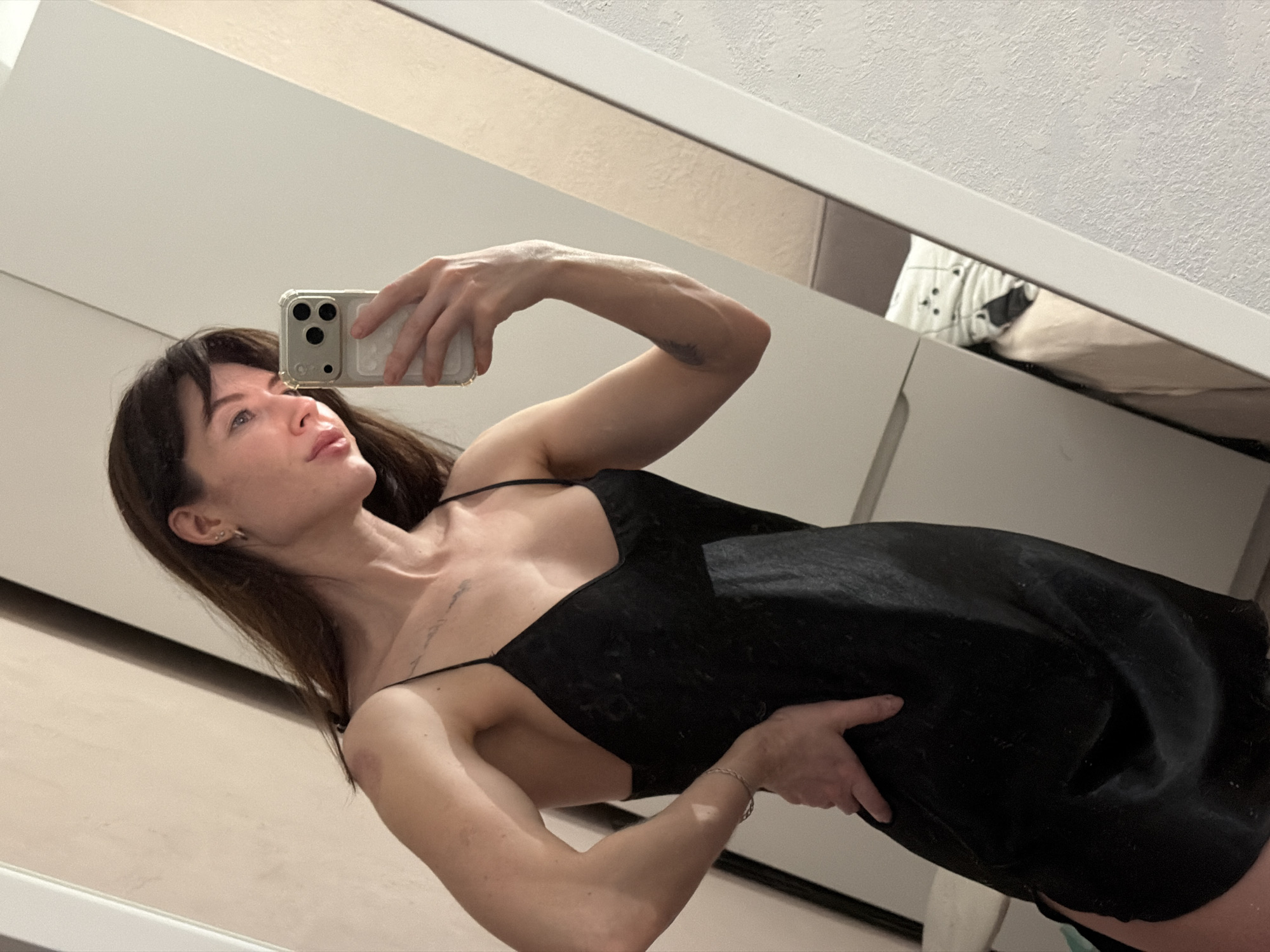 A woman is taking a selfie in a mirror. She has long brown hair and is wearing a black dress with spaghetti straps. She is holding her phone with her right hand and has her left hand on her hip. The mirror is reflecting a white wall and a bed. The exact location in Perm, Russia cannot be determined from the provided image.