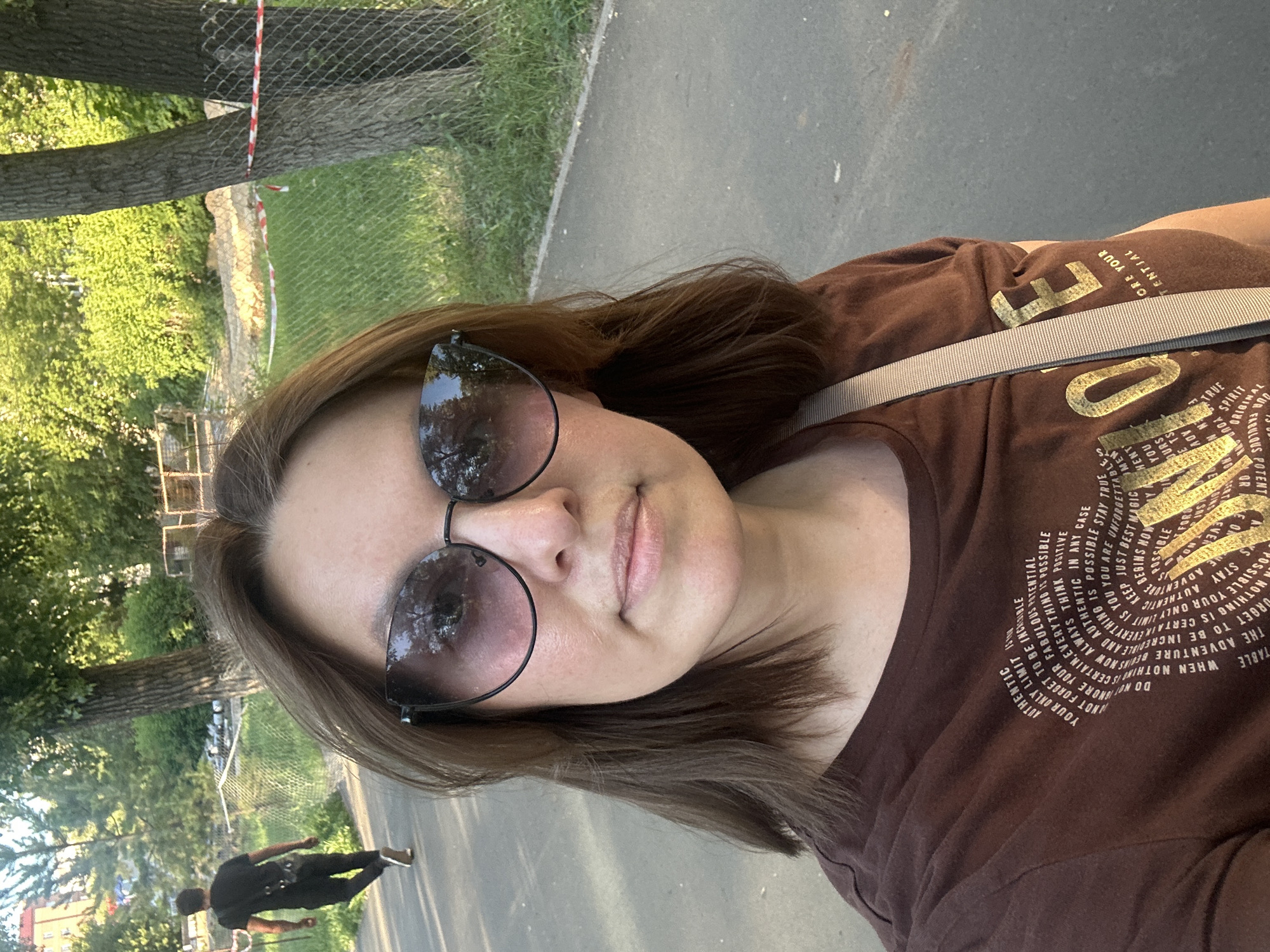 A medium shot captures a woman taking a selfie outdoors on a sunny day. She has medium-length brown hair and is wearing large, cat-eye sunglasses. Her expression is neutral, and she is looking directly at the camera. She is wearing a brown t-shirt with gold text printed on it, and a light-colored strap from a bag or purse is visible across her shoulder.
The background shows a paved pathway curving to the right. To the left of the path, a man in a black t-shirt and pants is walking away from the camera. Beyond him, there is a green grassy area with trees and some construction fencing with red and white tape. The location is identified as Orenburg, Russia, suggesting a park or urban green space.