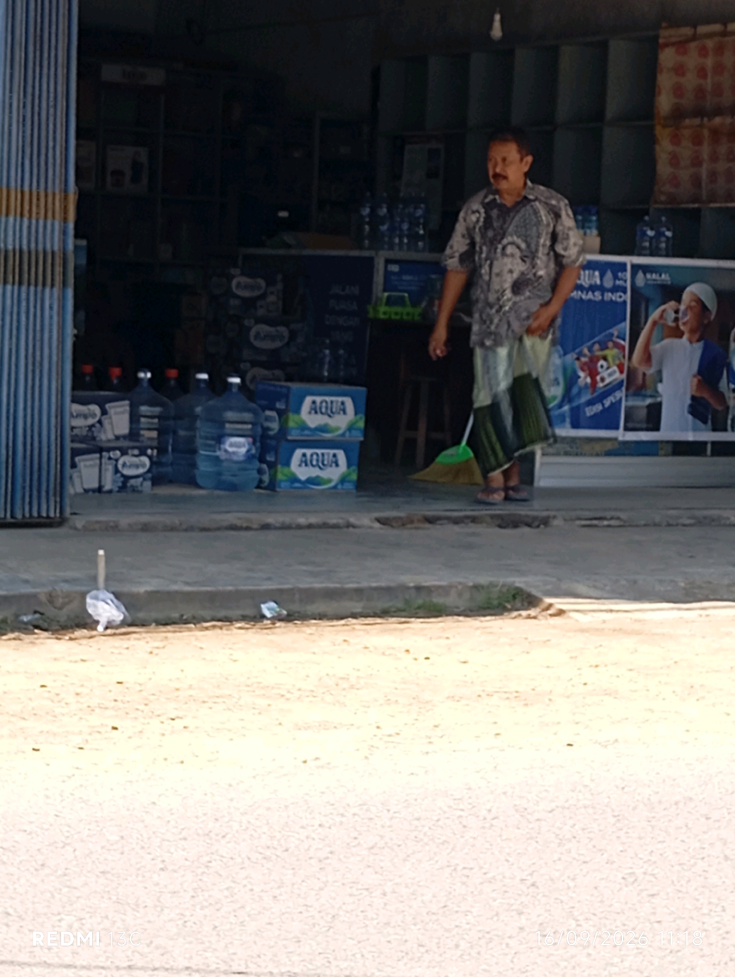 In Wuluhan, Indonesia, a man stands outside a shop, holding a broom. He is wearing a patterned shirt and a sarong. Behind him, several large water jugs are stacked, and a blue sign displays images of people and text. To the right, a young boy in a white shirt drinks from a bottle. The shop entrance is partly obscured by a blue corrugated metal shutter. The ground in front of the shop is paved, with some debris visible.