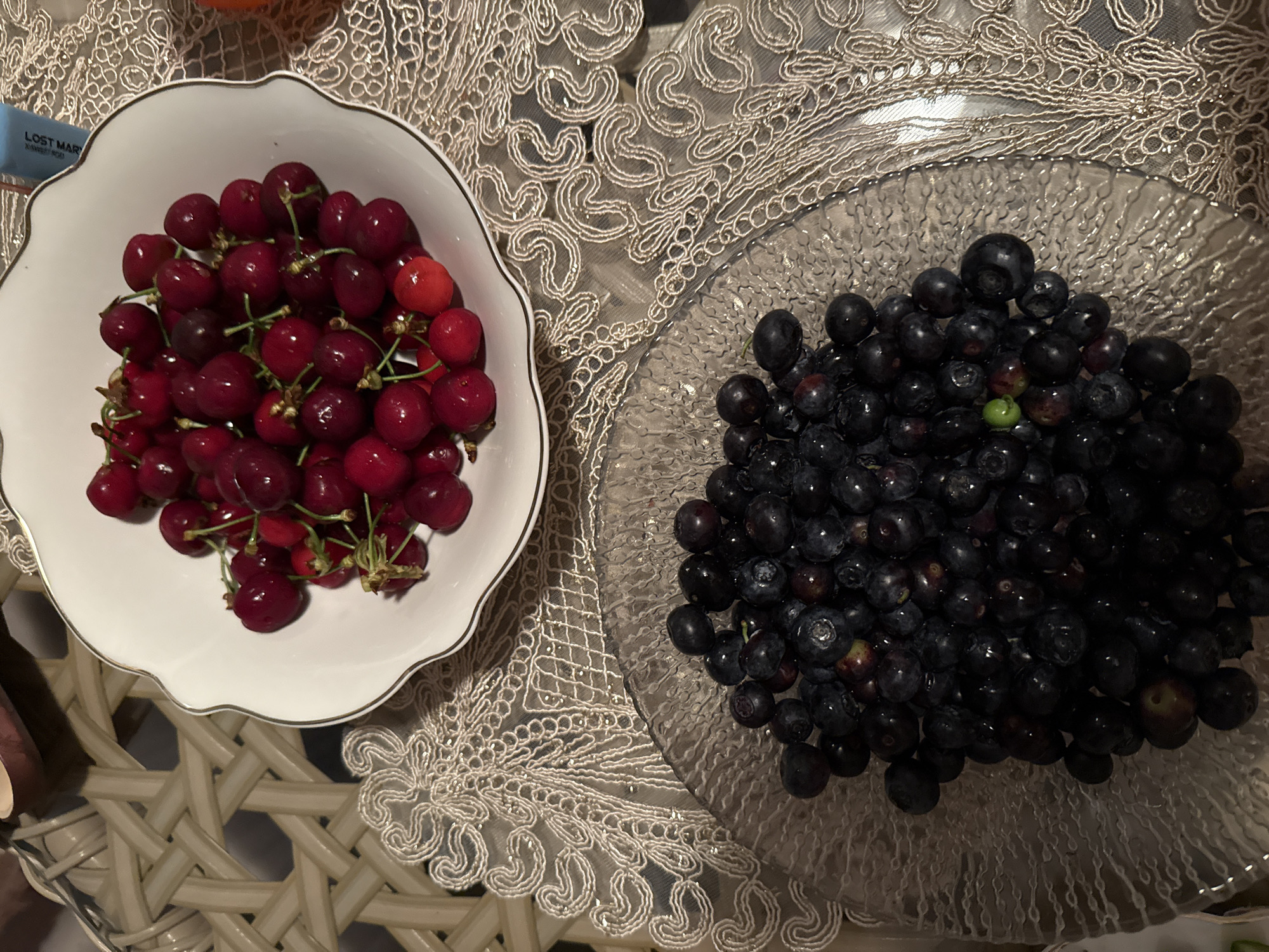 A top-down shot shows a bowl of cherries and a plate of blueberries on a table covered with a lace tablecloth. The bowl with cherries is white and has a decorative gold rim, while the blueberries are arranged in a clear glass plate. The background reveals a wicker table. The location appears to be indoors.