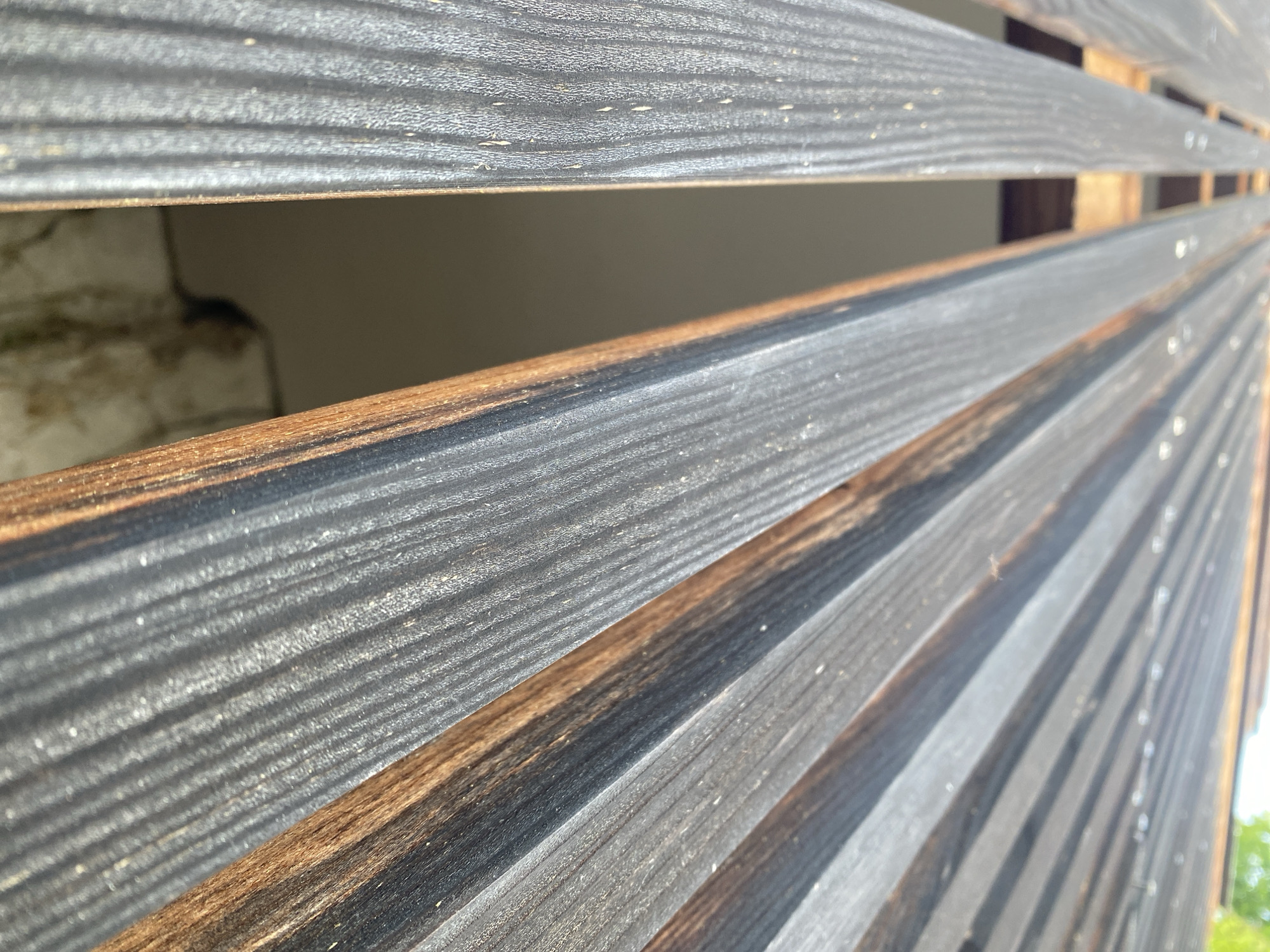 The image is a close-up, low-angle shot of weathered wooden planks, likely part of a deck or walkway, in Hallstatt, Austria. The planks are dark gray with visible wood grain and some reddish-brown undertones, suggesting they may be charred or treated. They are laid parallel to each other with narrow gaps between them. Sunlight illuminates the scene from the upper right, casting shadows and highlighting the texture of the wood. Through the gaps, a dimly lit interior space is partially visible, with what appears to be a textured wall. No people or animals are present. The overall impression is one of rustic, outdoor architecture.