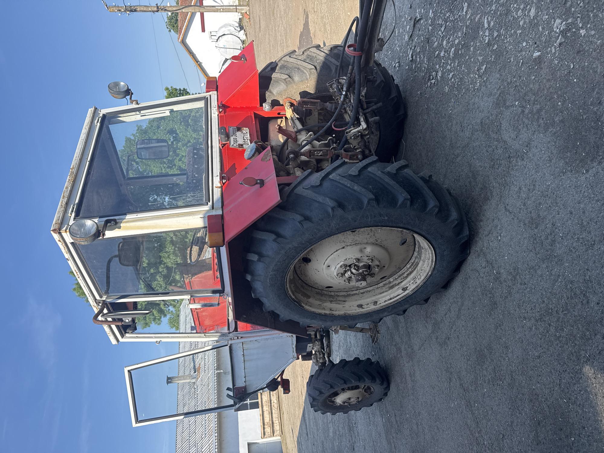 The image shows a red tractor parked on a concrete surface in Sarnevo, Bulgaria. The tractor has a white cab with an open door. The rear tires are large and black, while the front tire is smaller. A license plate reading "СТАРА ЗАГОРА 0157 КГ" is visible on the tractor. In the background, there are buildings and a clear blue sky. There are no people visible in the frame.