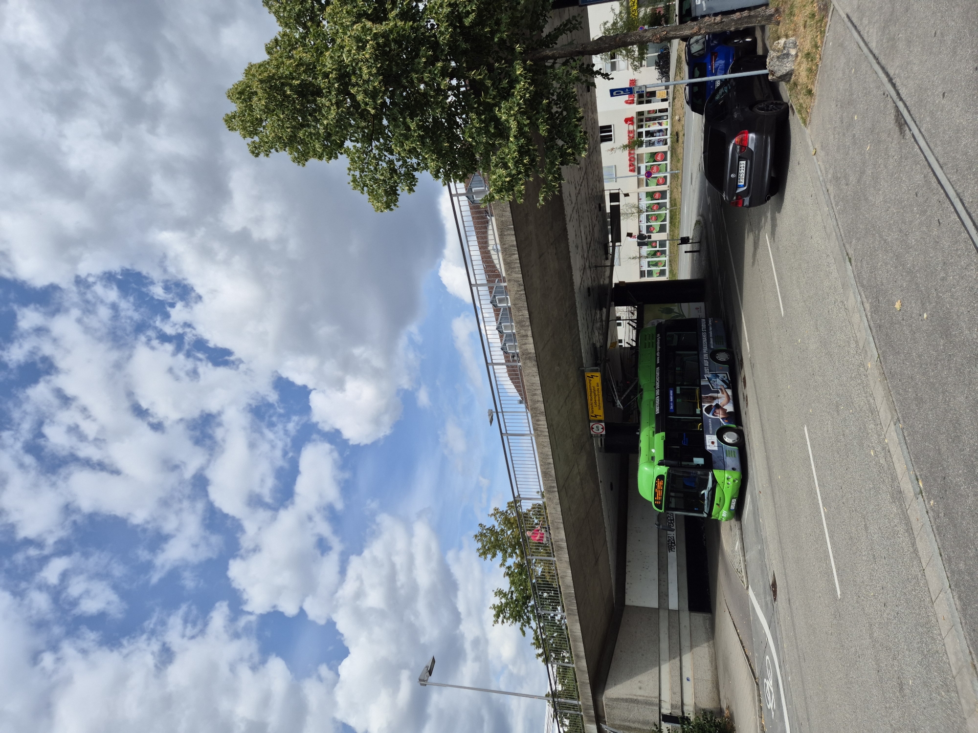 In Regensburg, Germany, a bright green trolleybus with advertisements for "Lust auf ein Praxis-Studium" is positioned under a concrete overpass. The bus, identified by license plate R-GV 904, is on a road that curves to the left. To the right of the bus, a dark gray BMW sedan with license plate R-CS 113 and a dark blue SUV are parked, partially obscured by a tree. In the background, a row of buildings includes "APOTHEKE" signage. The sky above is filled with scattered white clouds against a bright blue backdrop. The scene is captured from a slightly elevated perspective, looking upwards towards the overpass.