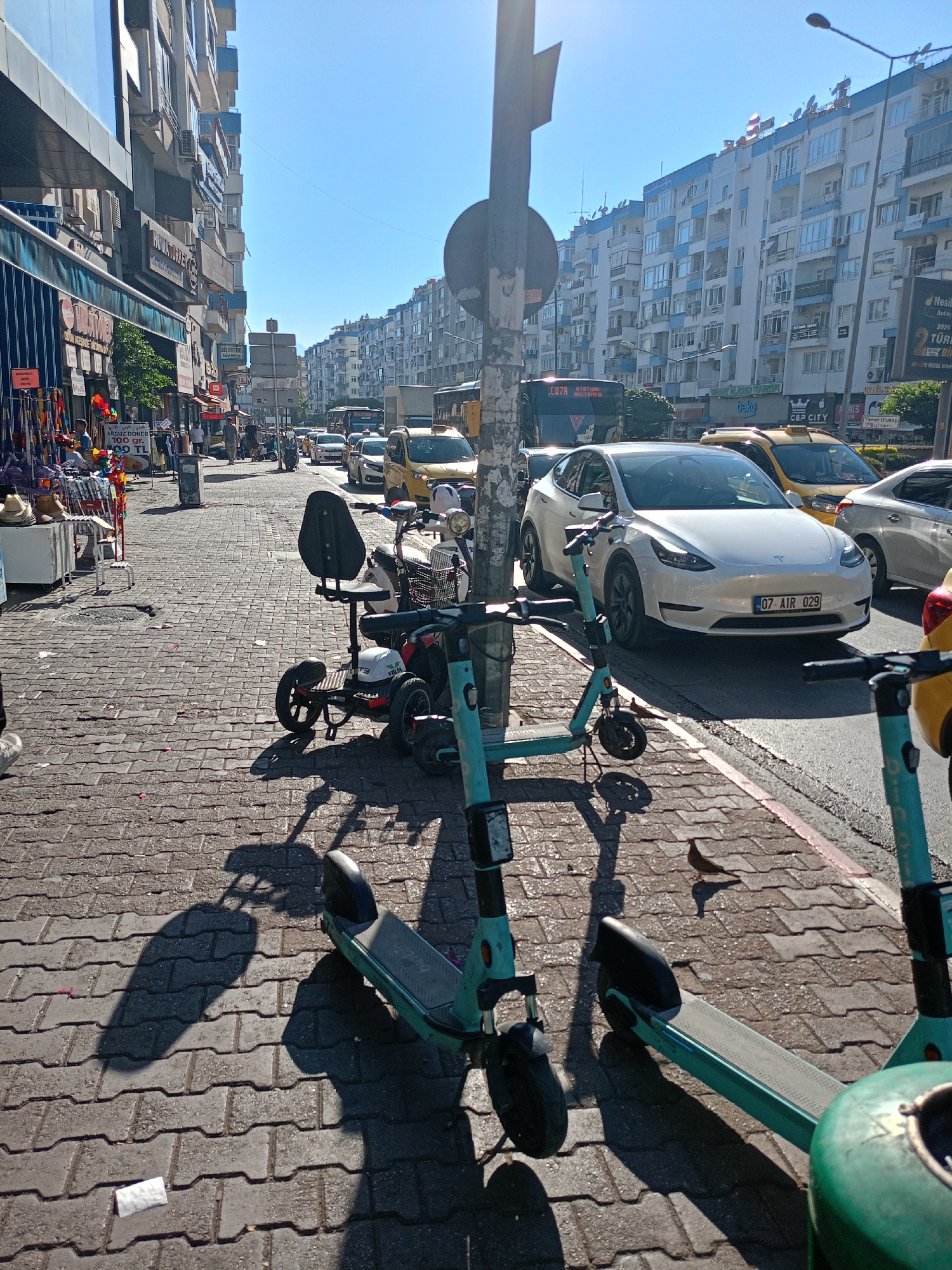 The image depicts a street scene in Antalya, Turkey, likely in a commercial area with multi-story buildings lining the street. The foreground is dominated by a cobblestone sidewalk where several parked electric scooters are visible. One scooter in the immediate foreground is teal-colored and partially cropped out. To its left, another teal scooter is parked, casting a long shadow. A three-wheeled electric mobility scooter with a seat and a basket is positioned further back on the sidewalk.
The street itself is busy with traffic. A white Tesla Model Y with license plate "07 AIR 029" is prominently featured, stopped in traffic. Behind it, a yellow taxi is visible, along with other vehicles including a bus and a silver car. The buildings on both sides of the street are modern apartment complexes with numerous balconies and windows. The sky is clear and blue, indicating a sunny day. On the left side of the street, there are storefronts with awnings and displays of merchandise, including clothing and colorful items. A few pedestrians are scattered on the sidewalk. A bird is perched on the sidewalk in the mid-ground.