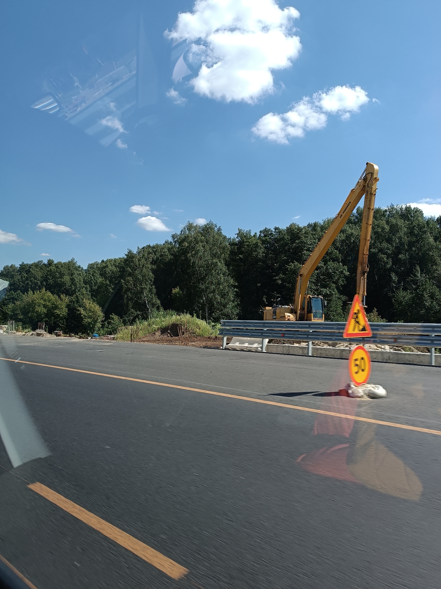 The image shows a construction site with an excavator, a guardrail, and traffic signs on a sunny day in Kraskovo, Russia. The excavator is yellow and appears to be parked. There are two traffic signs: a yellow triangular sign with a black image of a person working with a shovel, and a circular yellow sign with the number "50" in black. The road is paved and has orange lane markings. Trees line the background, and there are some clouds in the blue sky. The image is taken from a moving vehicle, as evidenced by the motion blur on the road and the reflection of what appears to be a car interior in the upper left portion of the image. No people are visible in the image.