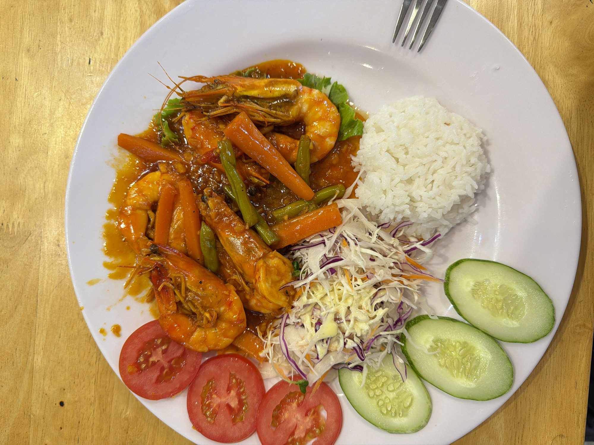 A top-down photograph displays a meal served on a white oval plate, situated on a light brown wooden table, consistent with a setting in Nha Trang Ward, Vietnam. The plate features a central portion of several cooked, shell-on shrimp, heads and tails intact, thoroughly coated in a glossy orange-red sauce. This main component includes elongated, tapered pieces of cooked carrots and thin green beans, resting on a base of green lettuce leaves. To the left of the shrimp, three round slices of red tomato are arranged above three oval slices of green cucumber. Adjacent to these, a mound of shredded salad is present, composed predominantly of finely shredded white cabbage, mixed with purple shredded cabbage and thin orange strands, topped with a small quantity of light-colored dressing. A compact dome of white steamed rice occupies the lower right section of the plate. A silver metal fork with four tines is partially visible on the far right edge of the plate. The wooden table exhibits visible grain and minor dark specks on its surface. No individuals are present in the image.