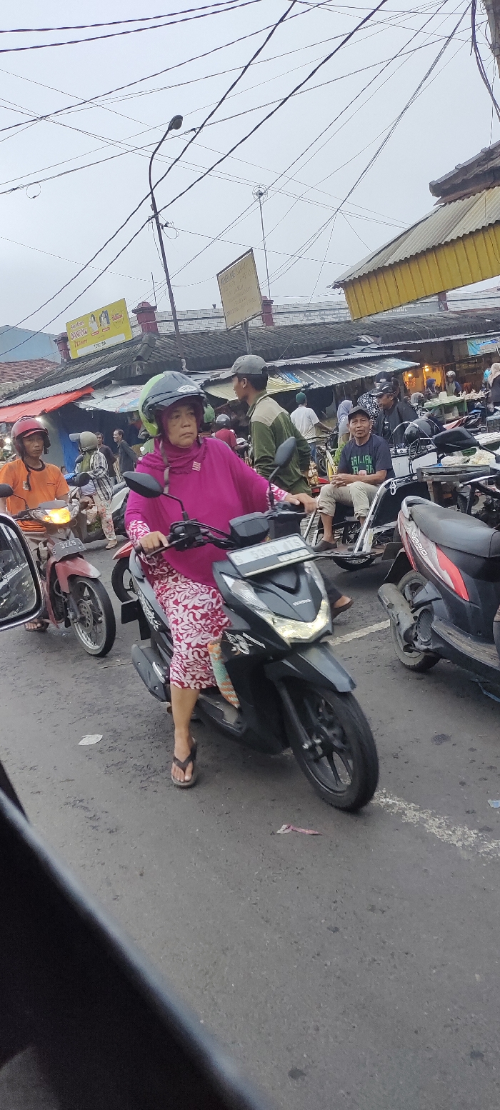 The image shows a street scene in Mojokerto, Indonesia (latitude: -7.468, longitude: 112.438). A woman wearing a pink hijab and a floral patterned dress is riding a black motorcycle. She is wearing a helmet and has a white scarf covering her hair. The motorcycle is stopped in traffic, and the woman is looking straight ahead. There are several other people in the background, including a man wearing a green shirt, a man wearing a black shirt, and a man wearing a blue shirt. There are several motorcycles parked in the background. The street is paved with asphalt and there are several power lines overhead. The sky is cloudy. The scene is busy and there is a lot of activity. The weather appears to be overcast. The scene appears to be in an urban setting. The woman appears to be in her 30s or 40s. The men in the background appear to be in their 20s or 30s. The image was taken from the perspective of a person in the street looking at the woman riding the motorcycle.