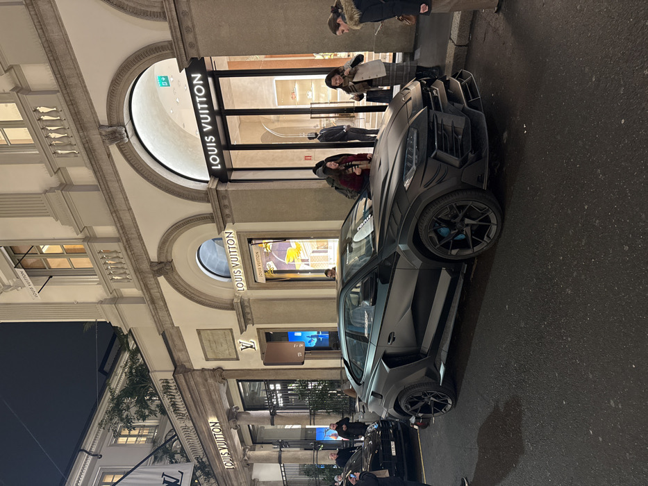 A matte black Mansory Lamborghini Urus parked on a street near a Louis Vuitton store. People mill about the sidewalk, some looking at the car and others entering the store. The scene is illuminated by storefront lights, casting a glow on the vehicle and the street. The building's architecture is ornate, with archways and classical details.