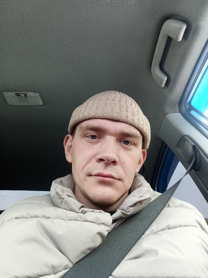 A single adult male is depicted in a front-facing portrait within the interior of a vehicle, located in Ufa, Russia. The individual, appearing to be in his twenties or thirties, has light skin, light-colored eyes, and short facial stubble. He is wearing a light beige ribbed knit beanie and a light beige or cream puffer jacket. A dark grey seatbelt is fastened across his chest and left shoulder. The vehicle's interior, visible in the background, includes a grey headliner, an overhead light fixture positioned to the top left, and a grey plastic grab handle located on the upper right side. A section of a blue vehicle frame or pillar is visible on the right. Through the windows, a bright blue sky is discernible on the right, while a blurry, light-colored exterior environment is visible on the left. The individual maintains a neutral expression, looking directly at the camera.