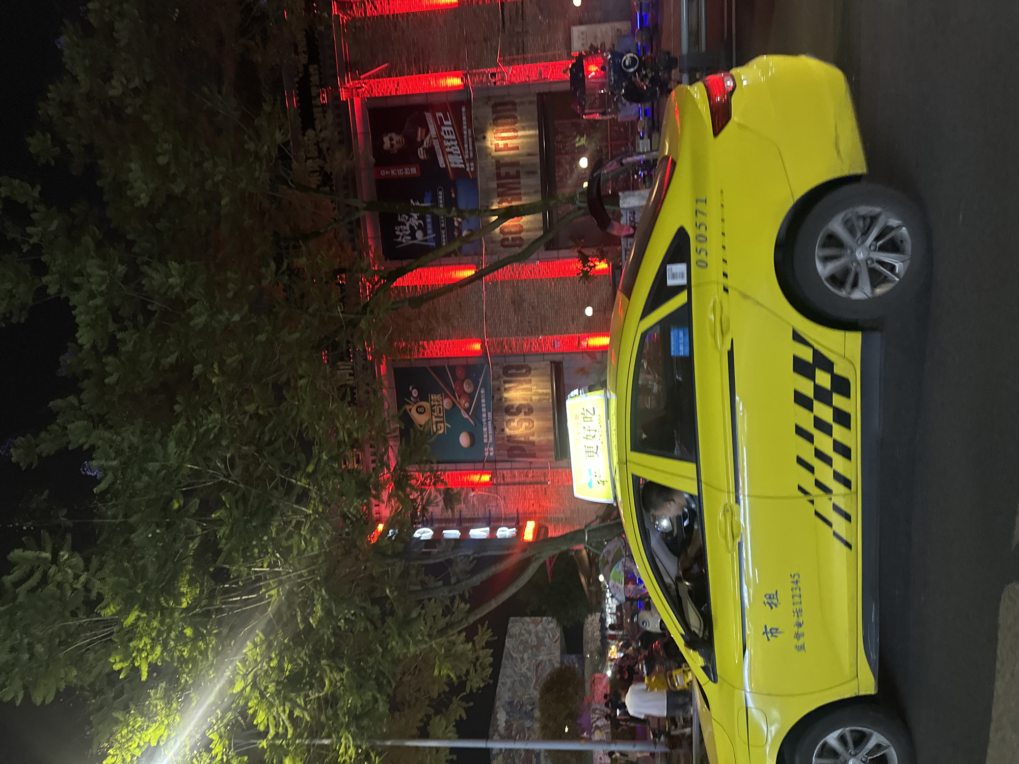 A yellow taxi is driving down a street at night. The taxi has a lit sign on its roof with Chinese characters. The building behind the taxi is decorated with red lights and has posters on its facade. The street is lined with trees. There are people and motorcycles on the sidewalk. The taxi has a license plate with the number "050571" and a checkerboard pattern on its side. The building in the background appears to be a commercial establishment.