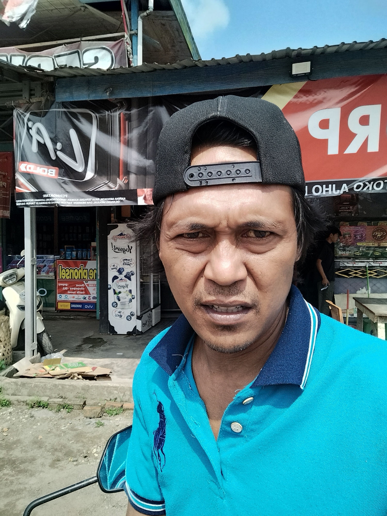 A man wearing a blue polo shirt and a black baseball cap is taking a selfie. He has dark hair and a goatee. He is standing in front of a store with banners and advertisements. There is a white scooter to his left and a wooden table to his right. The scene appears to be outdoors, possibly in a market or a street in Serdang Jaya, Indonesia. The banners show text in what appears to be Indonesian. One banner has the text "RP" in large white letters on a red background. Another banner, seen in a crop, displays a logo with the letters "V.J" and the word "BOROL" below it in white text on a black background. Further to the left, another banner reads "leznonger en" in large yellow text. A white refrigerator-like appliance with a cow graphic is visible to the man's right, displaying what appears to be yogurt packaging. A man in a dark shirt can be seen in the background, standing inside the store. The ground is paved with concrete and dirt. The sky is blue and clear.
