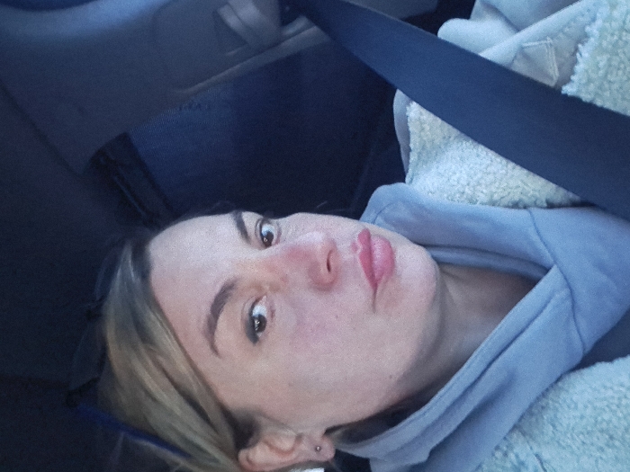 A woman is shown from a low angle, presumably in a vehicle, as indicated by the seat belt. She wears a light blue hoodie. The car interior and a fluffy white material are also visible. The woman has fair skin, light hair, and her lips are pursed in a possible pout. A small piercing is seen on her earlobe.