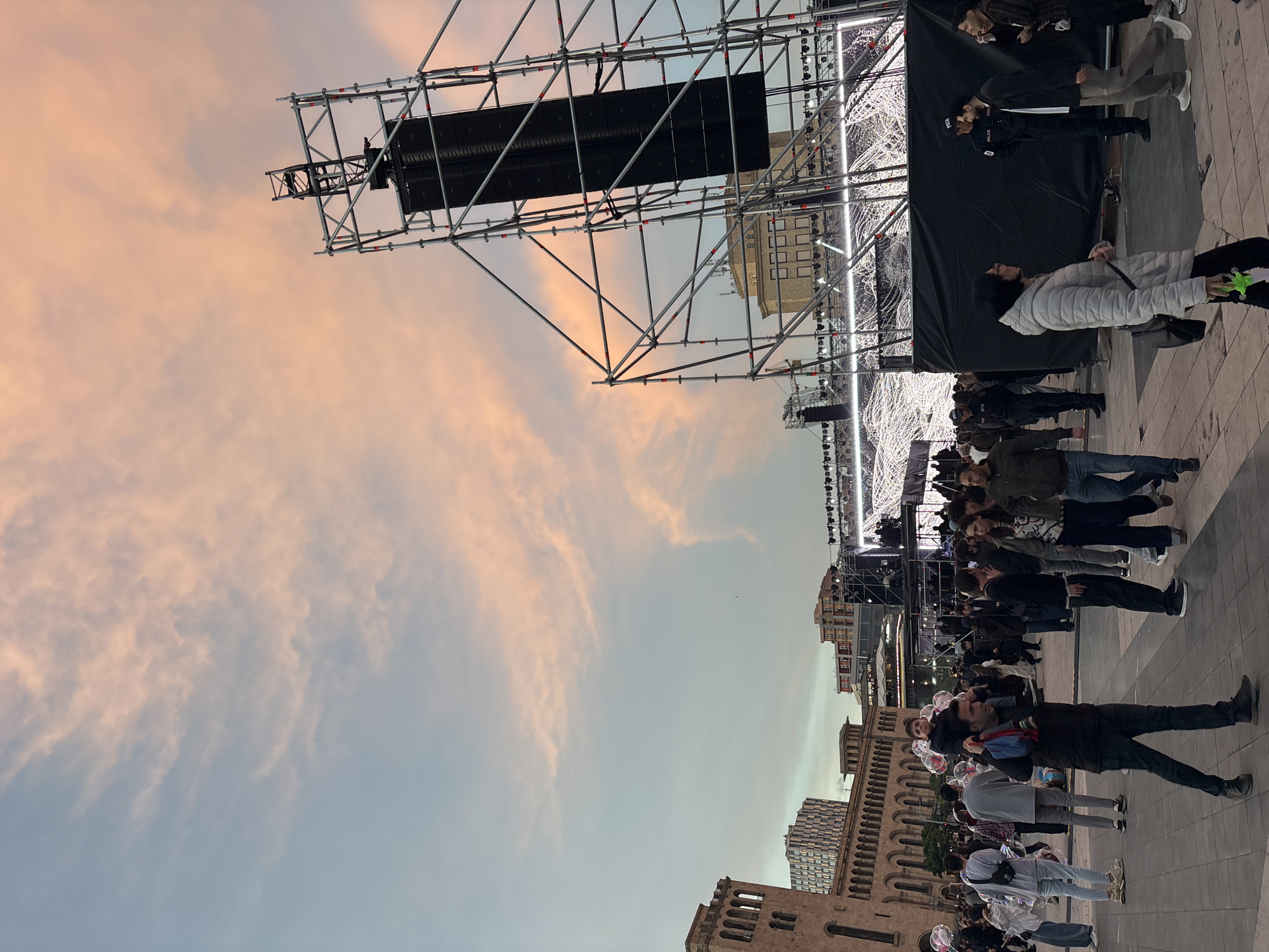 The image depicts a public gathering in Yerevan, Armenia. A stage structure, with scaffolding and what appears to be a sound system, stands in the background against a sky tinged with pink and orange hues. A crowd of people of varying ages and attire are present in the foreground. Some are standing still, while others are walking. Several buildings, including some with distinctive architectural features, can be seen in the cityscape. A police officer stands among the crowd.
