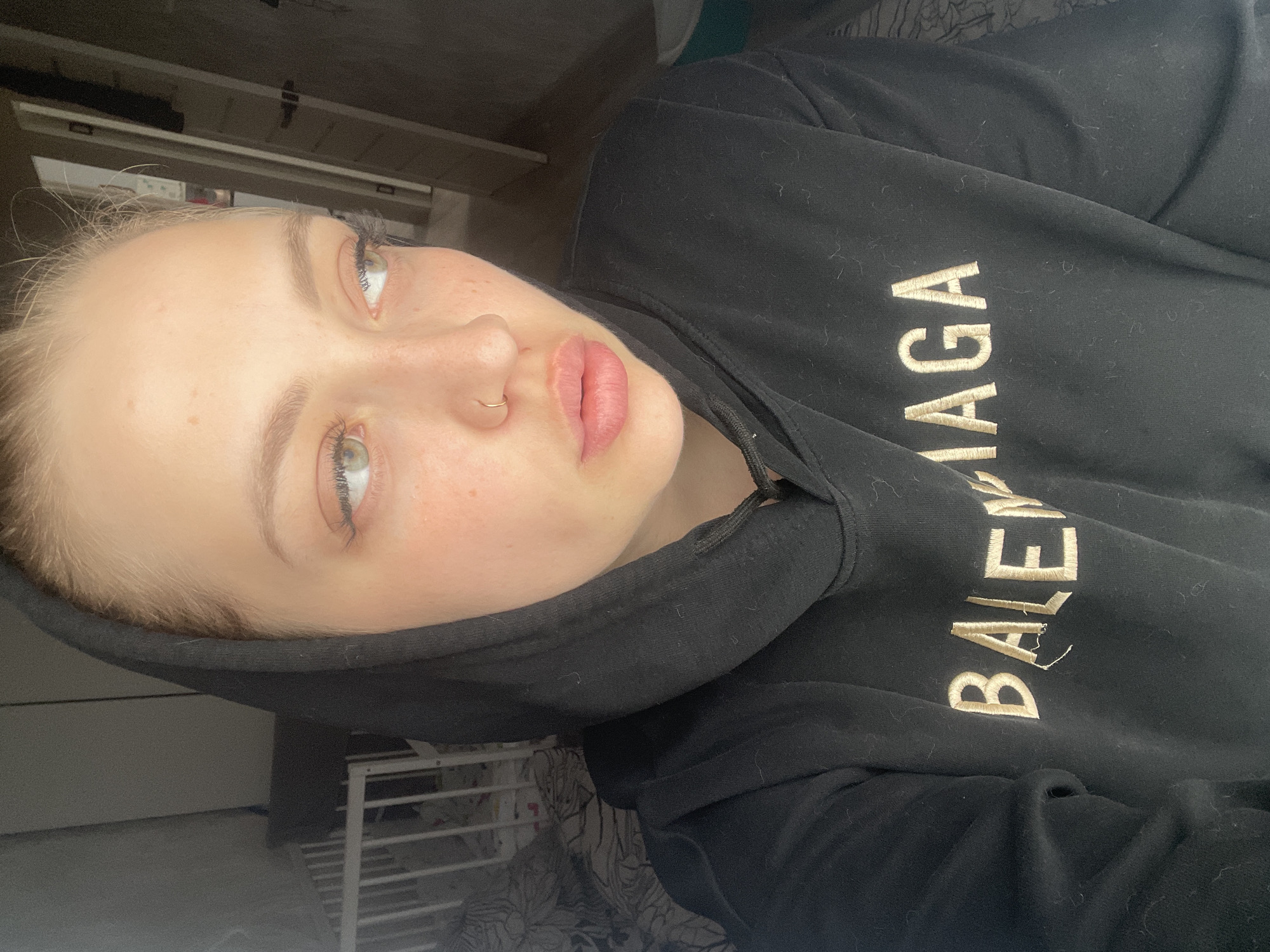 A young woman poses for a photo. She is wearing a black hoodie with "BALENCIAGA" embroidered on it. She has fair skin, light hair, green eyes, and a nose ring. The location is not apparent in the image. The photo appears to have been taken indoors.