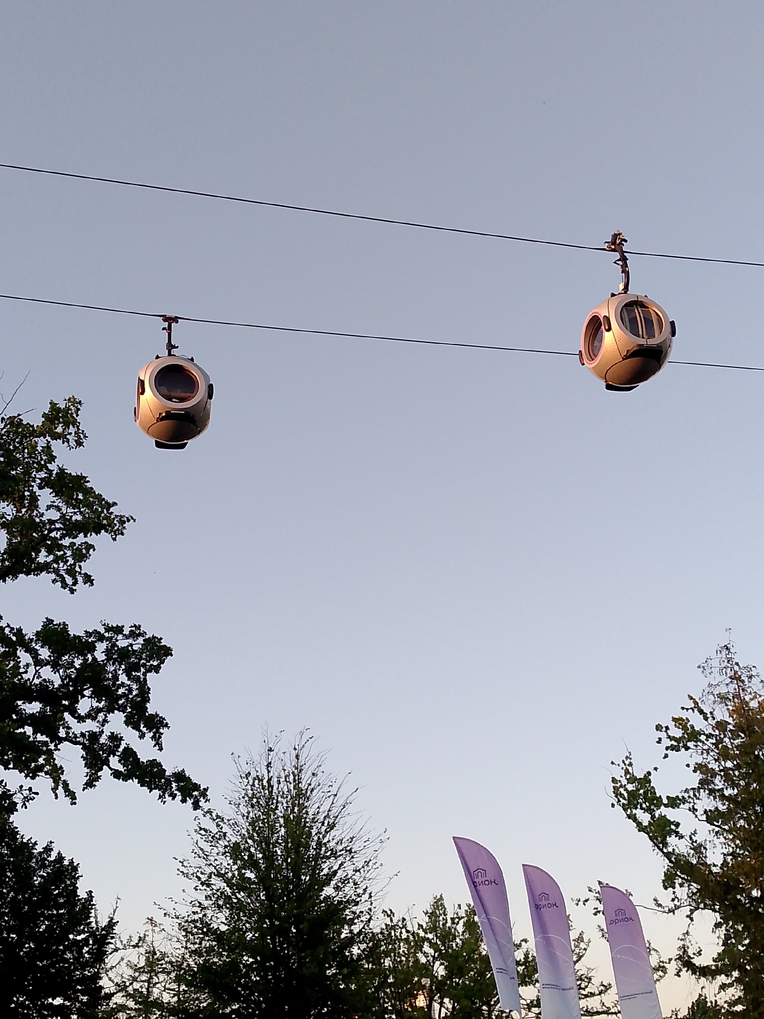 The image shows two cable cars suspended from a cable against a clear sky. The cable cars are spherical in shape with large windows. Trees are visible at the bottom of the image, silhouetted against the sky. Three purple flags with white text are also visible in the foreground. The scene appears to be an outdoor location, possibly a park or a tourist attraction. The presence of cable cars suggests a mountainous or elevated area. The flags may indicate an event or a specific location.