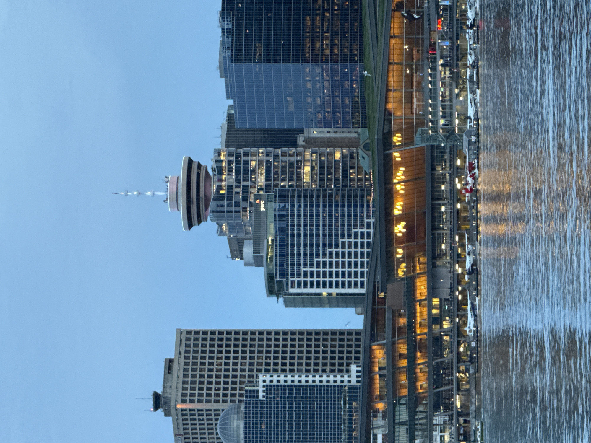 The image depicts the Vancouver, Canada, skyline at dusk, rotated 90 degrees counterclockwise. Dominating the left of the frame is the iconic Vancouver Lookout, a distinctive tower with a cylindrical base and a conical top. Several high-rise buildings fill the middle of the frame, featuring grid-like facades, predominantly in blue and white. To the right, warm yellow lights illuminate the waterfront area. The water's surface, reflecting the city lights, occupies the rightmost portion of the image, displaying a textured pattern. No people are discernible.