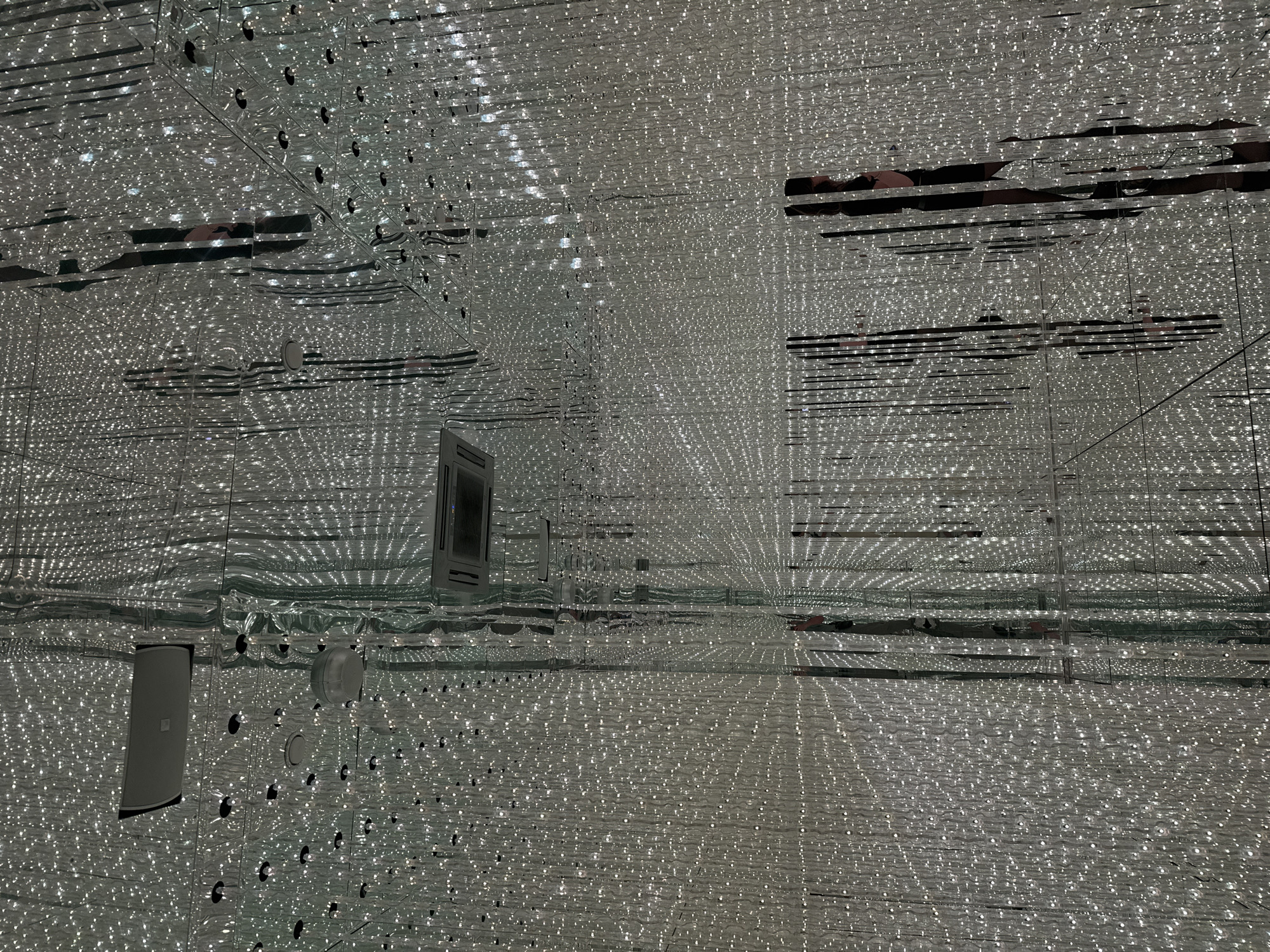 The image depicts an art installation, likely within the Museum of Illusions in Moscow, Russia. The scene is characterized by a mirrored room filled with countless small, bright lights that create an illusion of infinite depth. Several ceiling-mounted air conditioning units and lighting fixtures are visible. Reflections of people, appearing as dark silhouettes against the bright lights, are present throughout the space, suggesting visitors are interacting with the installation. The overall impression is one of a disorienting and visually striking environment.