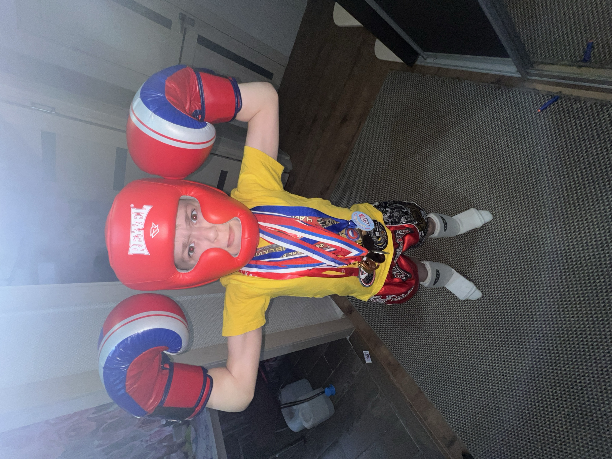 A boy is laying on a carpeted floor, wearing a red boxing helmet, red boxing gloves with blue and white stripes, a yellow t-shirt, and red shorts. He is also wearing white socks and has several medals around his neck on red, white, and blue ribbons. The text on one of the medals appears to include "СЛКВИ". There is a water bottle near his waist, and a door is visible in the background. Based on the text on one of the medals the boy is from Noviy, Russia.