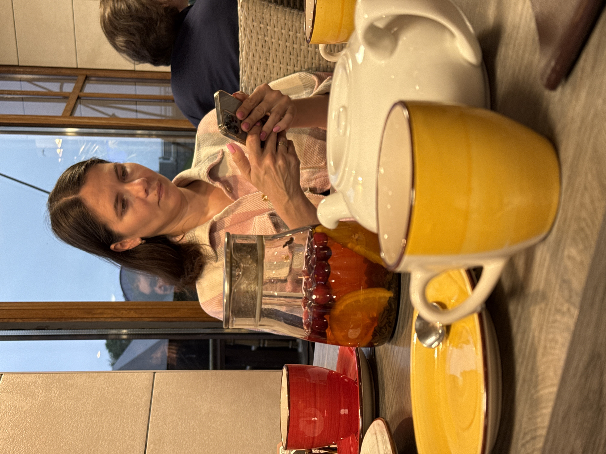 In Veliky Novgorod, Russia, a woman of Slavic descent is seated at a wooden table, engrossed in her smartphone. She wears a pink and white plaid shirt and has long, dark hair. Her hands, adorned with pink nail polish and a ring, hold the phone. In the foreground, a bright yellow mug and a yellow plate with a spoon are visible. To the left, a red teacup and saucer sit on the table. Behind the woman, a glass pitcher contains a beverage with fruit and orange slices. A white teapot is also on the table. The background reveals a window with a view of an evening sky and some greenery. Another person is partially visible to the right, seated in a wicker chair. The lighting suggests an indoor setting with natural light from the window.