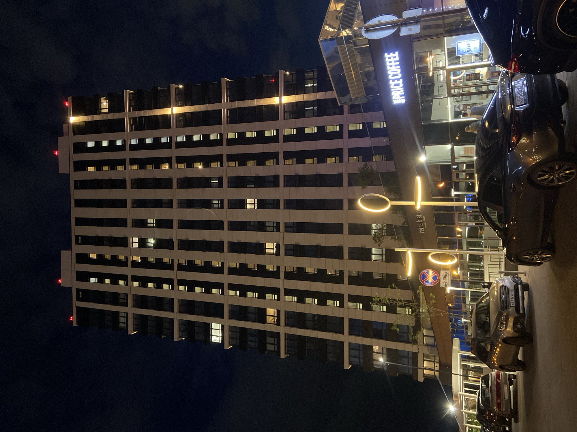 This image depicts an urban scene at night, confirmed to be in Kazan, Russia, by the presence of Russian license plates, specifically `К058ВХ 716` and `Р355ХМ 116` which correspond to the Republic of Tatarstan, where Kazan is the capital.
The central feature is a tall, modern, multi-story building, likely a residential or mixed-use structure, characterized by alternating vertical sections of dark glass and lighter, solid cladding. Numerous windows are illuminated with warm yellow light, indicating occupancy. Red obstruction lights are visible on the building's rooftop.
At street level, the building accommodates commercial establishments. A prominent, brightly illuminated sign for "PRICE COFFEE" is visible on the right, with a glass facade revealing two indistinct individuals inside. To the left of "PRICE COFFEE," a sign for "vétra bistro" is partially visible. The area in front of these establishments is a paved street or parking area, illuminated by modern streetlights featuring circular, glowing elements.
Several cars are parked or stopped in the foreground. From right to left: a dark-colored sedan (partially visible), a dark grey Mazda sedan with license plate `К058ВХ 716`, a silver Subaru Outback SUV with license plate `Х671КХ 163` (Samara Oblast region code, neighboring Tatarstan), and an older model silver sedan with license plate `Р355ХМ 116`. Another dark vehicle is partially visible on the far left.
Road signs are present on a pole near the "vétra bistro" entrance, including a red circular "No Stopping" sign (diagonal cross) and a "No Parking" sign (single diagonal line). The sky is dark blue with some subtle cloud formations. The overall impression is a vibrant, well-lit urban environment during nighttime hours.