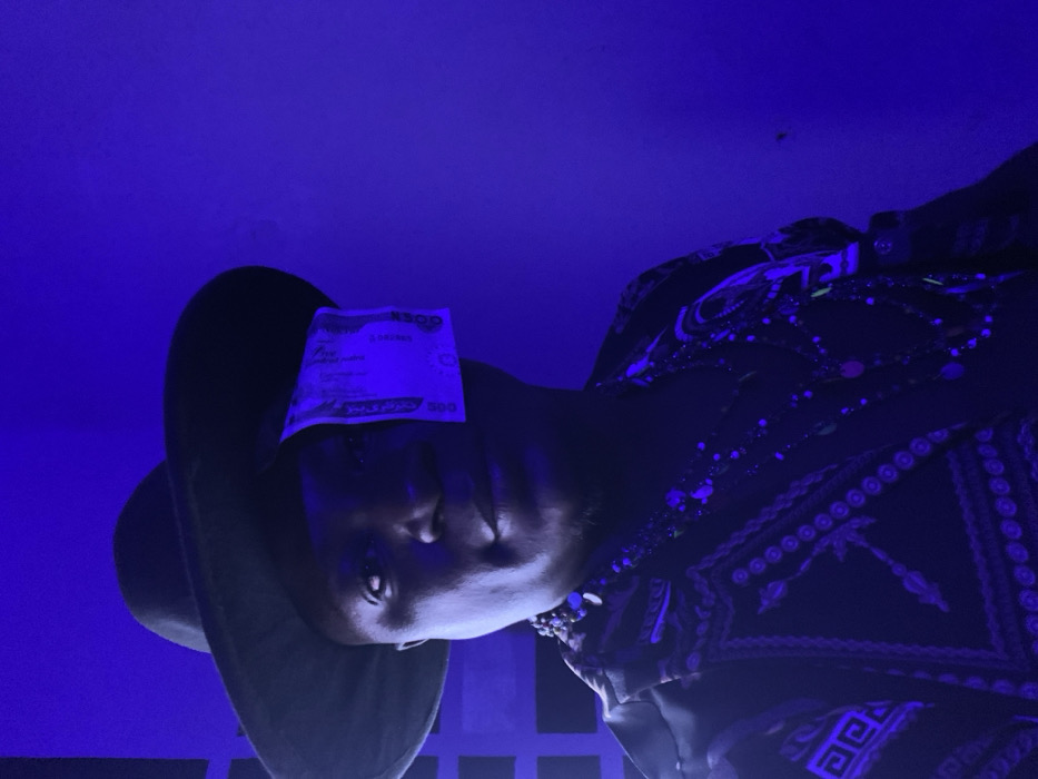 A person wearing a large hat has a 500 Nigerian naira note resting on their forehead, partially obscuring their face. The individual is dressed in a dark patterned shirt with jewelry visible around their neck. The lighting is predominantly blue, creating a dark and moody atmosphere. The image is rotated, presenting a slightly angled view of the subject. The background appears to be a simple wall.