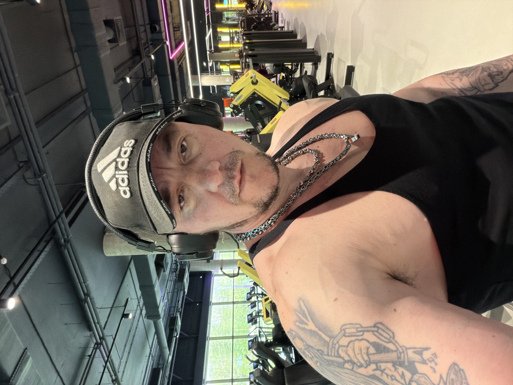 A man wearing a black tank top, grey Adidas baseball cap, and headphones poses for a selfie inside a gym. He has tattoos on his arm. Exercise machines are visible in the background. The location is not discernible as Moscow, Russia.