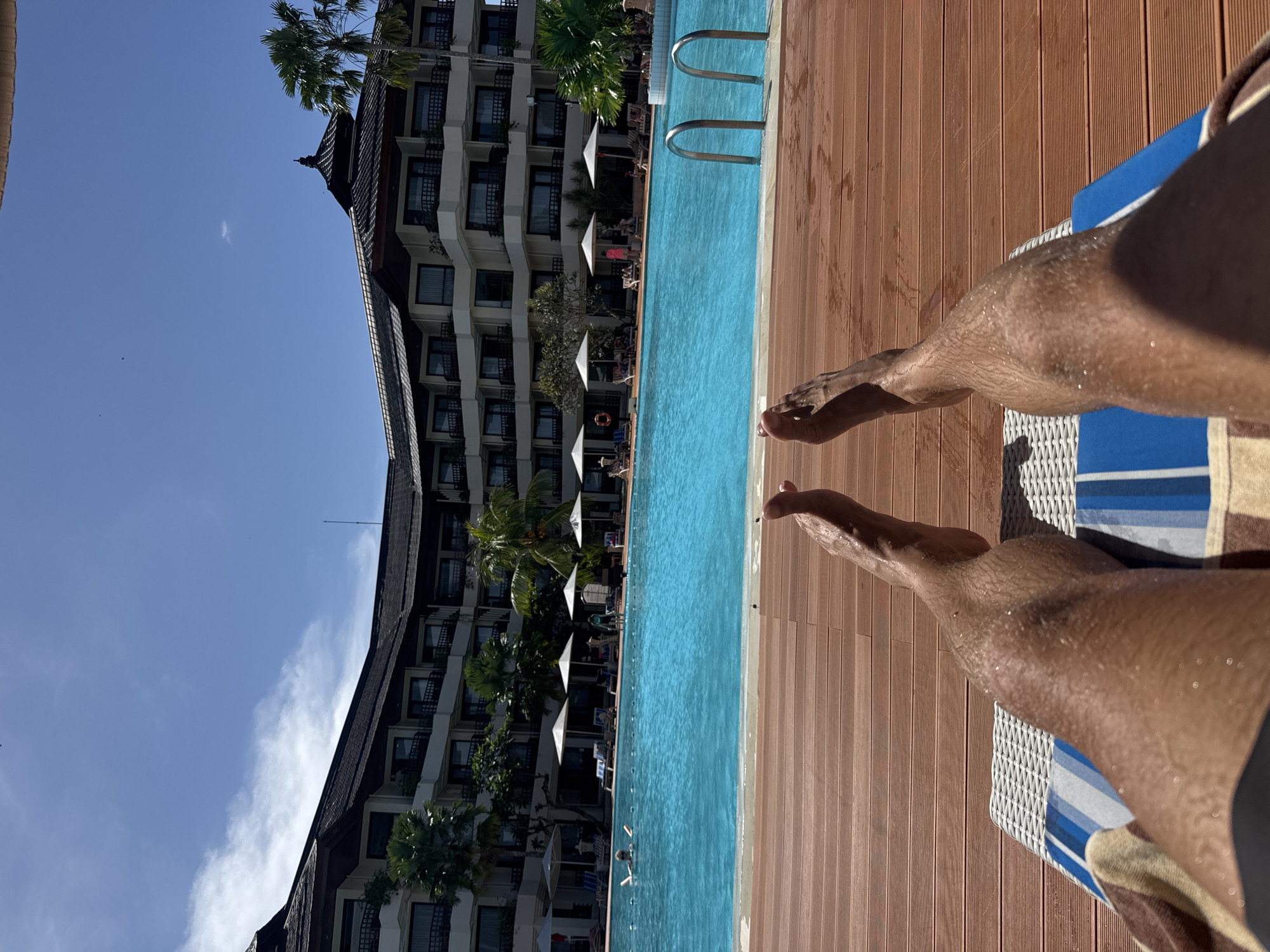 The image, taken from a first-person perspective and oriented horizontally, depicts a person relaxing by a swimming pool on a sunny day. In the foreground, tanned legs and feet, glistening with water droplets, are stretched out on a white wicker sun lounger, with a blue and white striped towel visible underneath.
The midground features a clear, turquoise blue swimming pool, bordered by a light brown wooden deck. Stainless steel pool ladders are visible on the far side of the pool. In the background, a large, multi-story hotel or resort building stands under a bright blue sky with a few wispy white clouds. The building showcases traditional dark rooflines and numerous windows or balconies.
Tropical palm trees and other greenery are interspersed around the building and along the pool's edge. Several white sun umbrellas and sun loungers are positioned beside the pool, with a few indistinct figures visible relaxing. One person is also seen in the pool. The scene suggests a tranquil, vacation-like setting at a resort during bright daylight, consistent with a tropical location like Denpasar, Indonesia. No visible text is present.