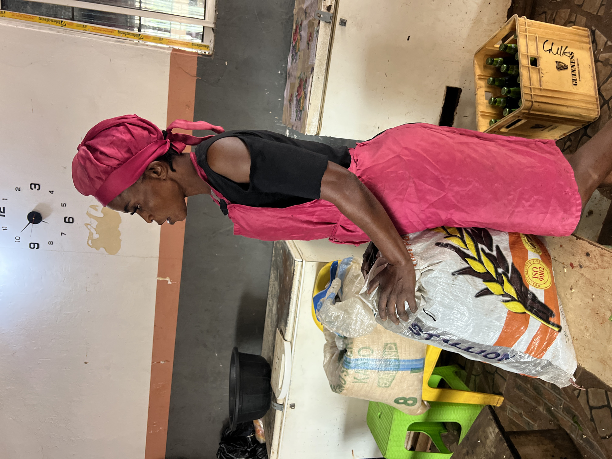 A person wearing a pink headwrap and a pink apron over a black t-shirt is carrying a large bag of what appears to be rice. The person is bending over, suggesting they are either lifting or moving the heavy bag. The scene is indoors, with a clock visible on the wall in the background showing approximately 9:15. There are also crates of Guinness beer bottles stacked in the background, and various plastic chairs are scattered around. The overall setting appears to be a storage area or a market stall. The lighting suggests it is daytime. The person's posture and the weight of the bag imply a task that requires effort. There is some peeling paint on the wall near the clock, and some of the items in the background are dusty, indicating a somewhat utilitarian or well-used environment. Text visible on the Guinness crate includes "Chilkes" and "Guinness". There is also text on the rice bag, with "GALLON" being partially legible.
