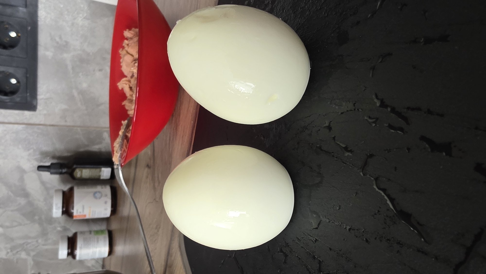 Two peeled hard-boiled eggs are placed on a black, textured surface with water droplets. In the background, a red bowl contains what appears to be shredded food, possibly tuna, and a fork. Behind the bowl are several bottles, including two pill bottles and a dark dropper bottle, situated on a countertop. The wall behind the countertop has a tiled texture. Electrical outlets are visible on the upper right portion of the wall. The scene appears to be in a kitchen environment.