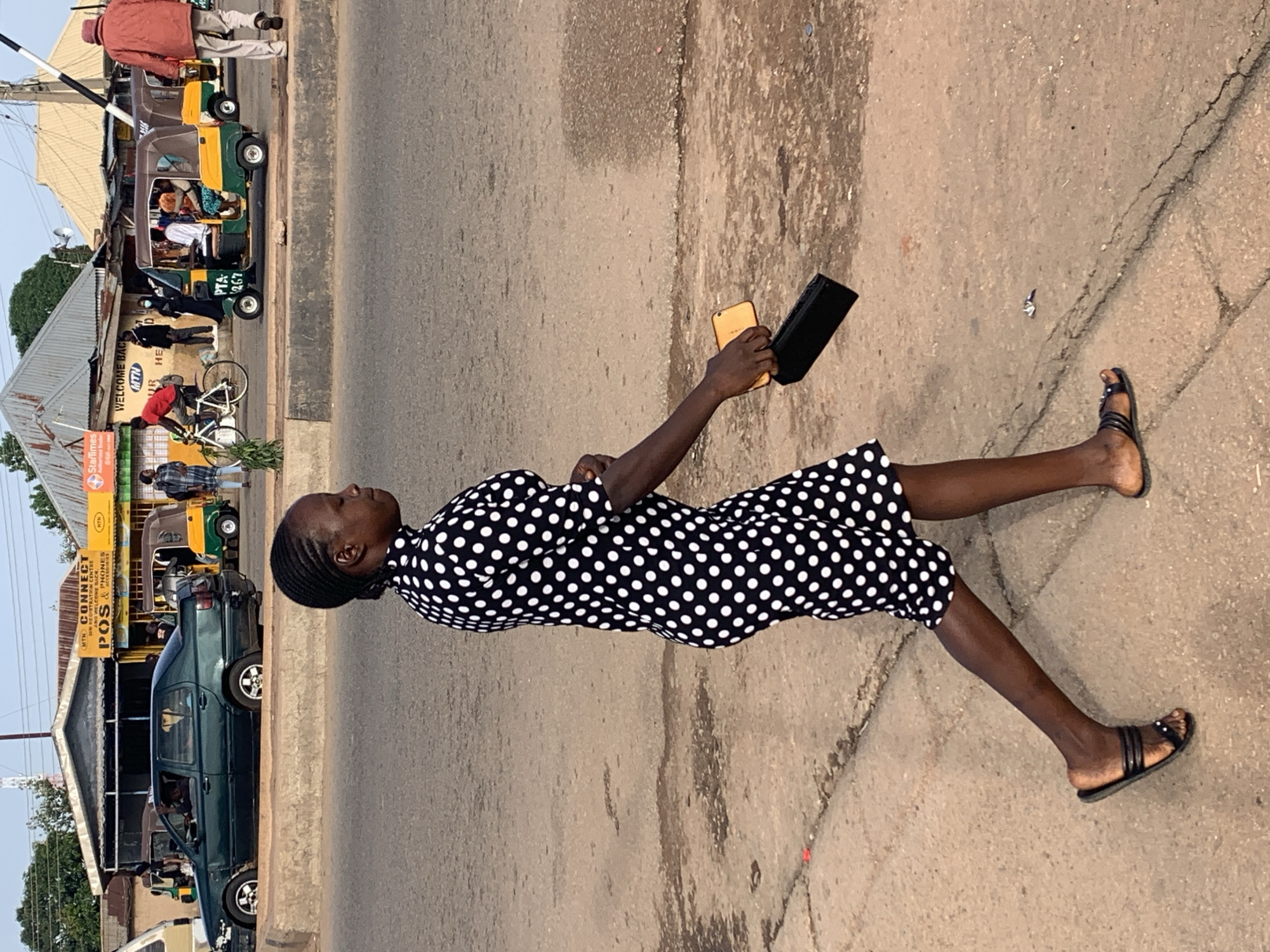 In Gura Top, Nigeria, a woman in a black and white polka dot dress walks down a paved street, holding a black clutch and a yellow cell phone. She wears black sandals. Behind her, a dark green car is parked on the side of the road, and a yellow and green motorized tricycle is visible further down the street, along with other parked vehicles and buildings. The scene appears to be a marketplace or a busy roadside commercial area, with signs for "MTN CONNECT CENTRE," "POS & PHONES," and "StarTimes" indicating various services available. The lighting suggests it is daytime. The woman's gaze is directed downwards, possibly at her phone, with a neutral expression. The overall atmosphere suggests a normal day in a bustling urban environment.