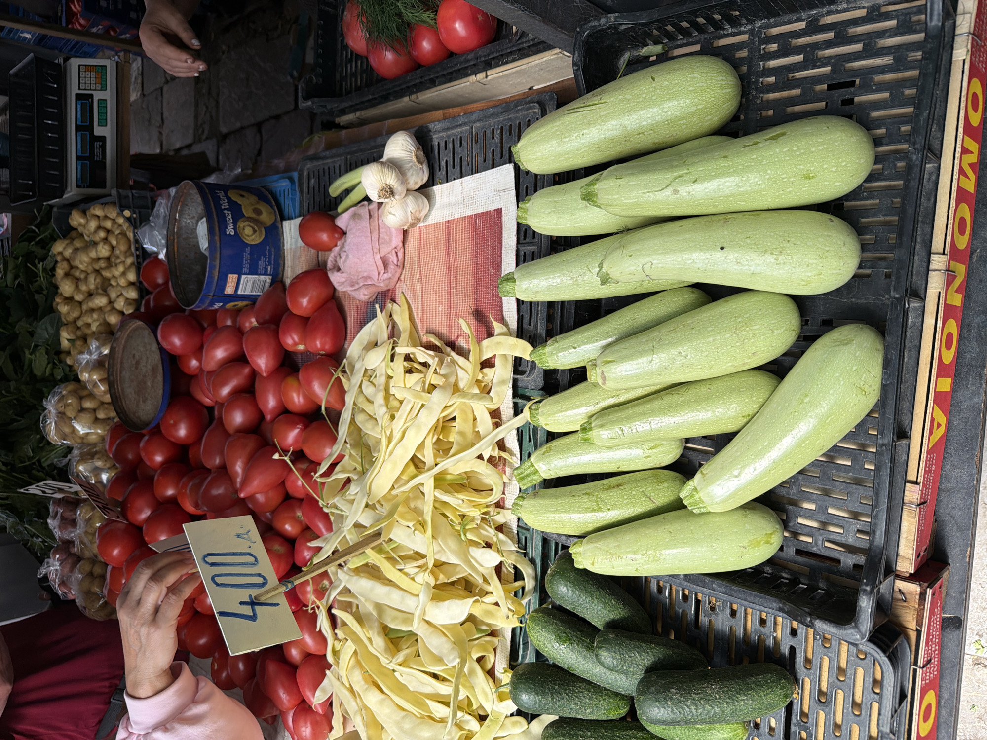 The image depicts a produce stall within a market environment in Vracar Urban Municipality, Serbia. The foreground features two black plastic crates. The crate on the left contains multiple dark green cucumbers of varying lengths. The crate on the right holds numerous light green courgettes (zucchini). Above these crates, a large quantity of pale yellow flat beans is displayed on a red and white striped fabric. Directly behind the flat beans is a substantial pile of red, elongated tomatoes. A small, rectangular, light yellow price tag, held by a wooden stick, is inserted into the tomatoes, displaying the handwritten numeral '400' followed by a period. A person's right hand, wearing a light pink sleeve, is positioned above these tomatoes. To the right of the central tomato pile, a crumpled pink cloth, a cluster of three garlic bulbs, and a single green onion stalk are visible. Further back, an aged blue metal tin labeled 'Sweet World' with images of biscuits is present. In the background, sacks of light brown potatoes and bundles of dark green leafy vegetables are displayed. On the far right, a digital weighing scale with blue displays is visible on a stand, and the hand of a second individual is partially seen near this area. Another black crate on the right contains more red tomatoes and green dill. The market floor appears to be paved.