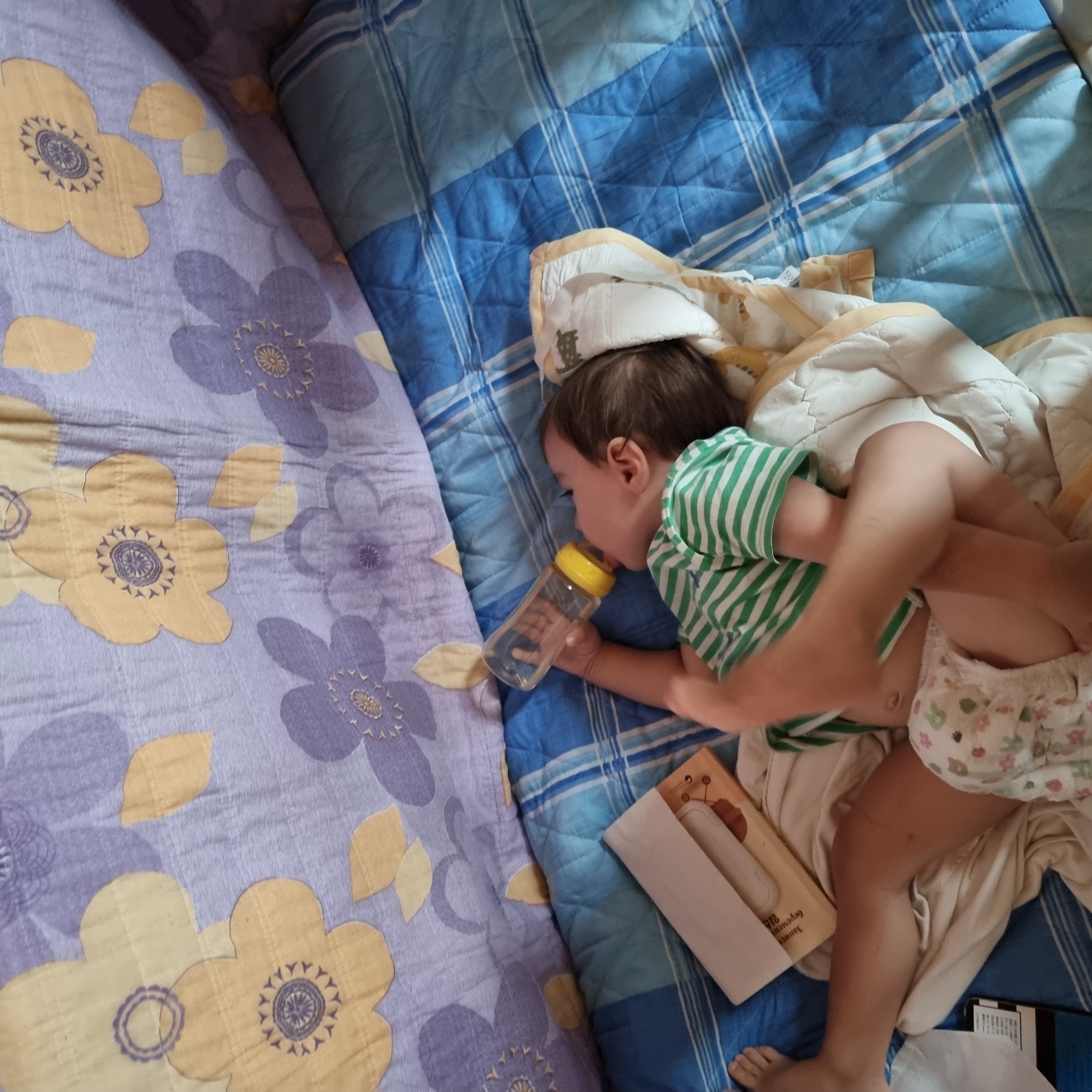 A baby is lying on their back, reaching for a baby bottle. The baby is wearing a green and white striped t-shirt and a diaper. The baby is on a blue and white checkered blanket. There is a pillow with a floral pattern behind the baby. The image appears to be taken indoors in a bedroom. This image does not contain any information about the city of Dunnae-myeon, South Korea.